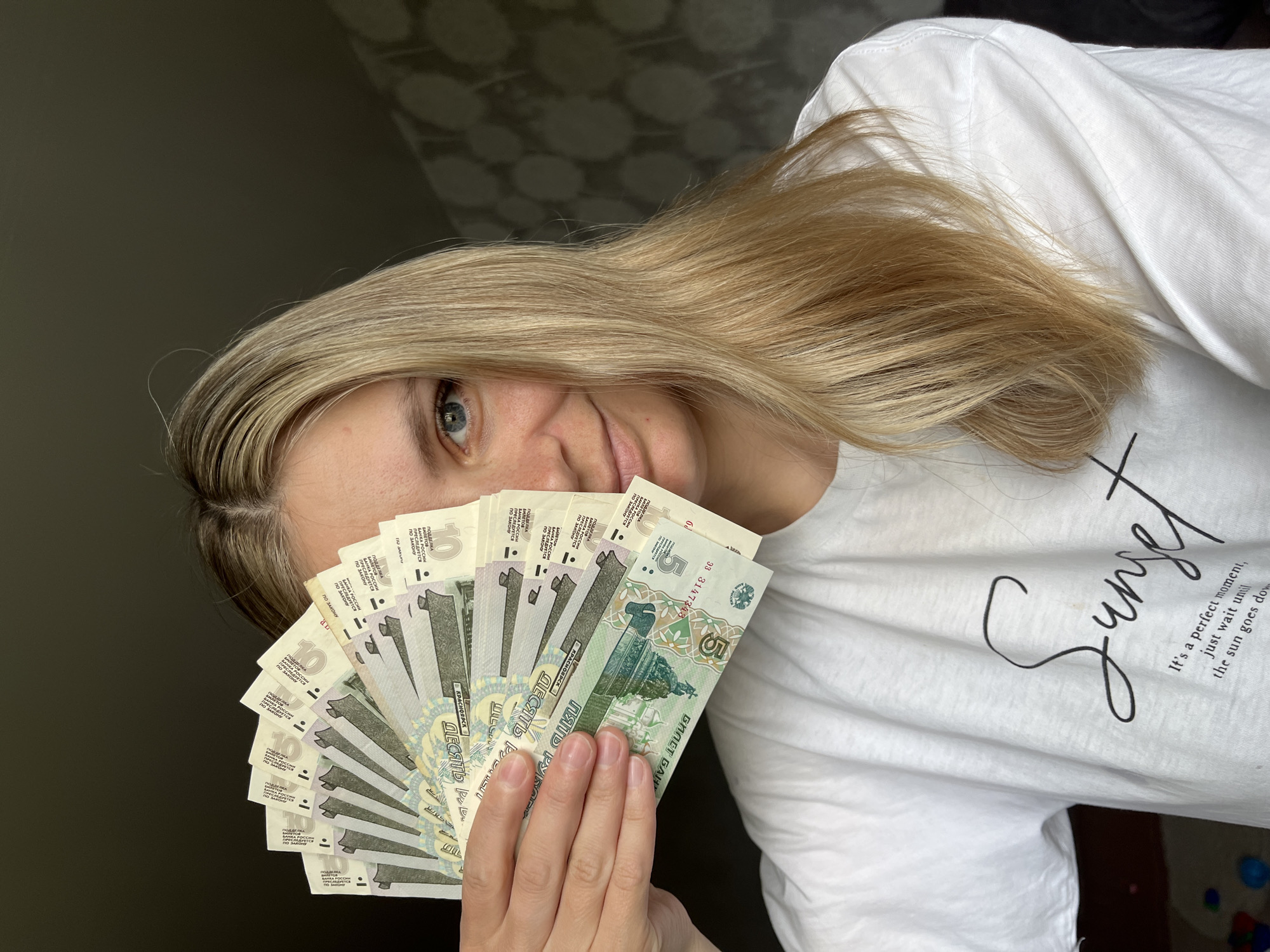 A fair-skinned woman with blonde hair partially covers her face with a fan of Russian banknotes. The banknotes are primarily 5 and 10 Ruble denominations. The woman wears a white t-shirt with the word "Sunset" printed on it, along with the phrase "It's a perfect moment, just wait until the sun goes down." The background is a dark, nondescript wall. The location in the city of Chaikovsky, Russia, cannot be determined from the image.