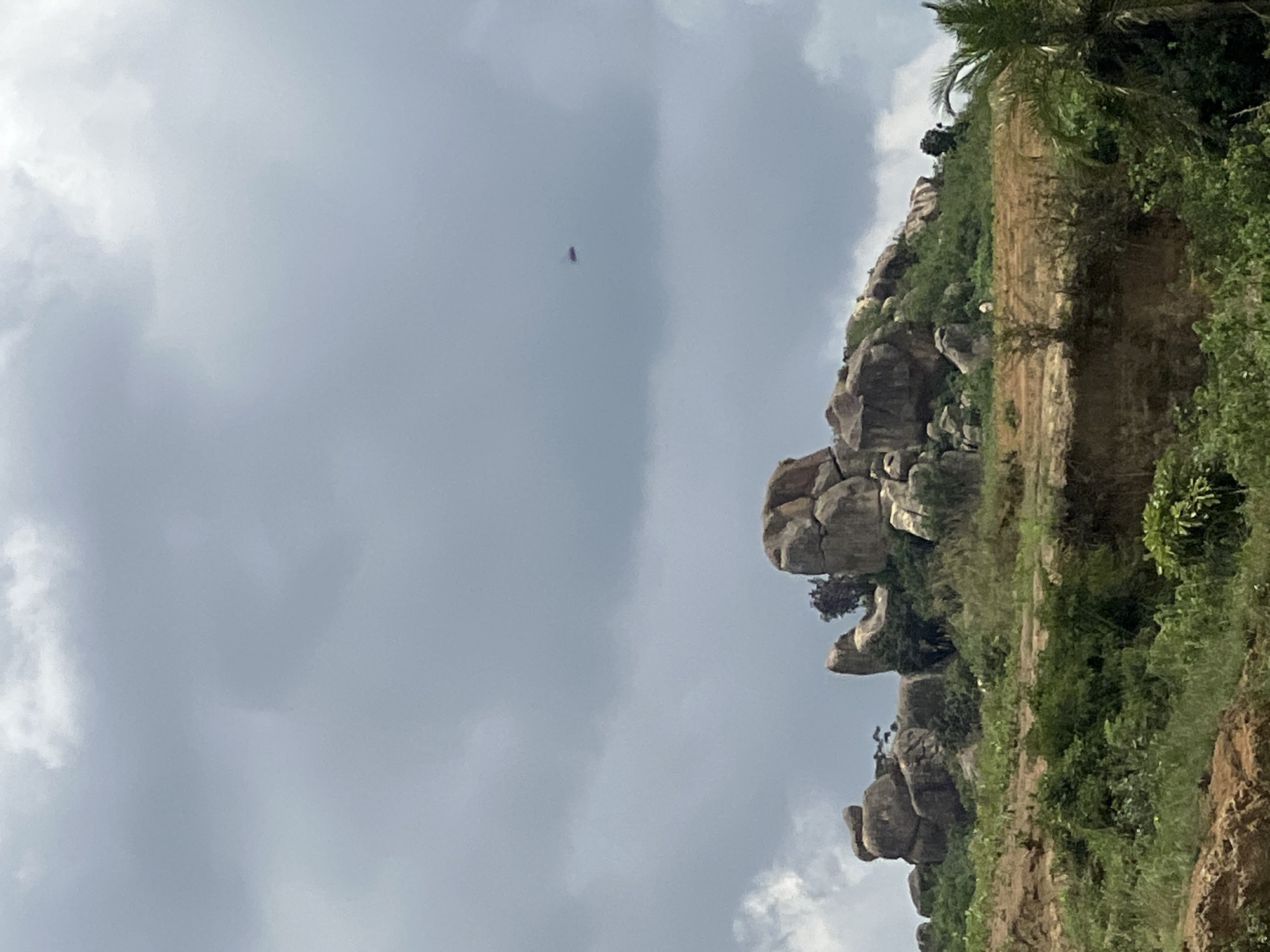 The image depicts a landscape in Jos, Nigeria, characterized by a rocky outcrop in the upper middle ground, topped with large, weathered boulders. The terrain leading up to the rocks is covered in lush green vegetation and appears to be somewhat cleared or terraced in places, with exposed brown earth. In the far distance, high in the cloudy sky, a small, reddish object, possibly a drone or a distant aircraft, is visible. The sky itself is overcast with dramatic grey clouds. The foreground shows more dense green foliage and a rough, earthy bank. The overall scene suggests a natural, somewhat rugged environment.