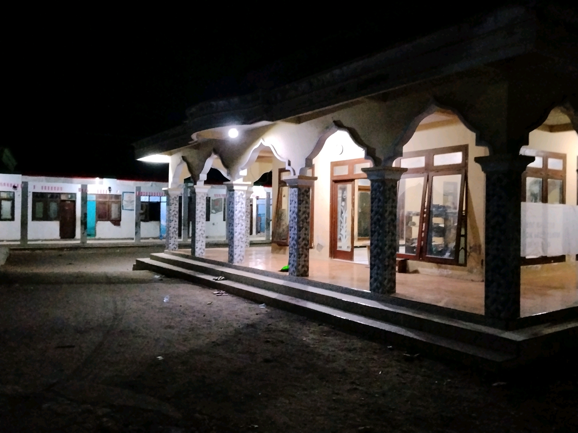 The image shows a dark, outdoor scene at night, likely in Panggungan, Indonesia. The focus is on a covered patio area of a building. The patio is supported by decorative columns with patterned tiles. Large windows and doors with wooden frames are visible along the back of the patio, and some are open. Inside the building, the floor appears to be tiled and polished, reflecting the light from an overhead lamp on the patio. The ground in front of the patio is dark and appears to be dirt or gravel. A set of concrete stairs leads up to the patio. A pair of flip-flops are visible on the second step from the bottom. The sky is black, indicating nighttime. In the background, another building with windows and what appear to be doors is visible, also illuminated. The overall scene suggests a residential or possibly a small commercial building in a rural or semi-rural setting.