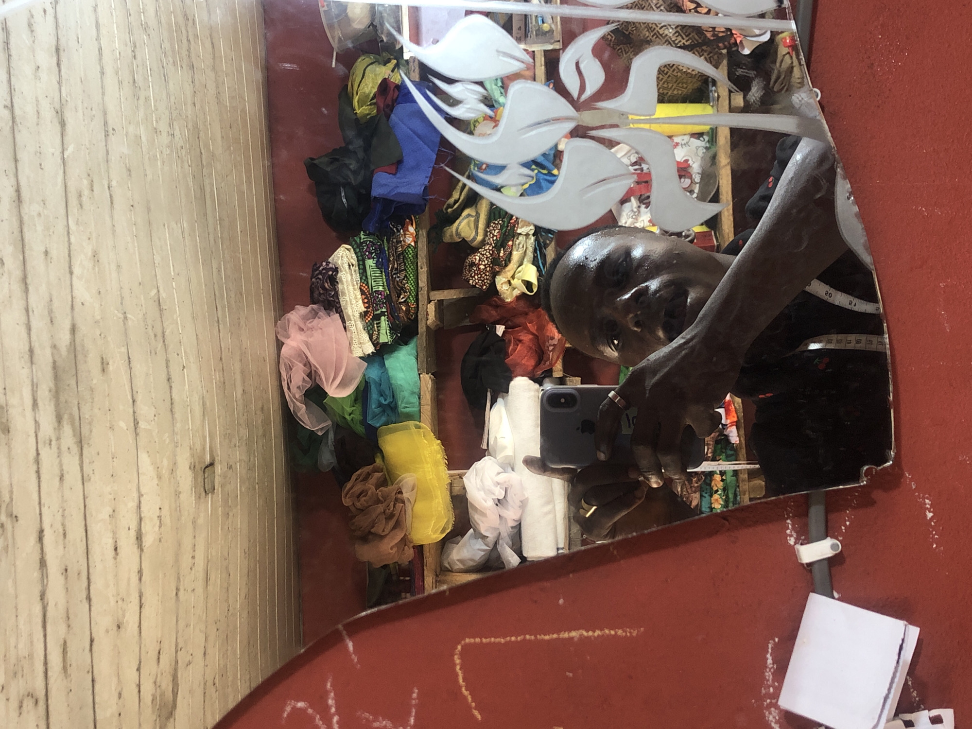 A man is taking a selfie in a mirror within a room in Ado, Nigeria. The room appears to be a workshop or storage area, with shelves filled with various fabrics, textiles, and possibly other sewing supplies. The man is wearing a black shirt and has a gold ring on his left hand and a silver ring on his right hand. He is holding a smartphone to take the picture. The mirror has a decorative etching of floral elements on its surface. The walls of the room are painted red. There is a piece of paper and a white clip attached to the red wall on the left side of the image. The ceiling is made of wooden planks.