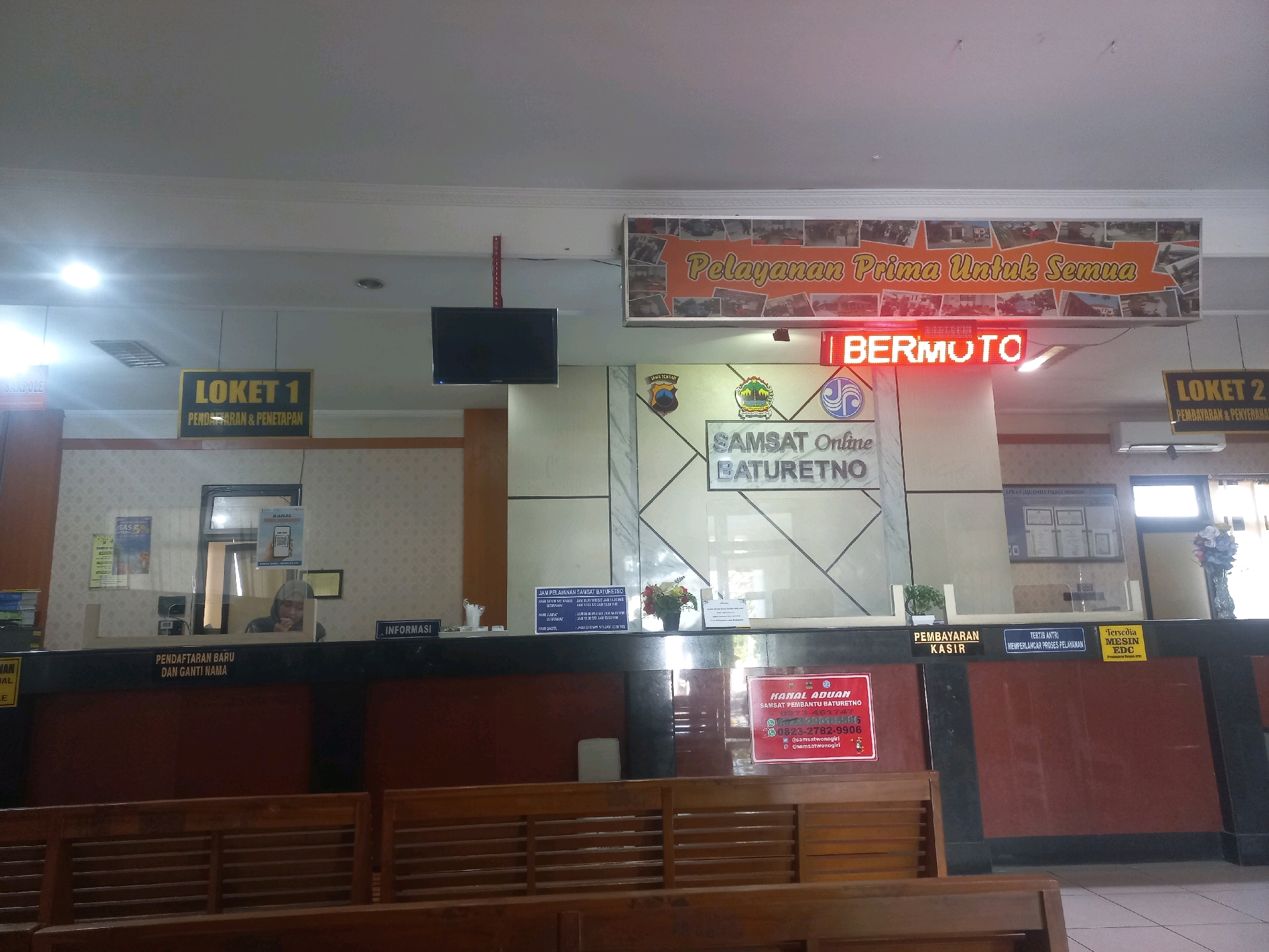 The image depicts the interior of a government service office, likely a SAM SATS (Sistem Administrasi Manunggal Satu Atap) or motor vehicle tax office, in Baturetno, Indonesia. The scene is a well-lit, public service counter area with wooden seating in the foreground.
Dominating the background is a large counter with multiple service windows. Above the counter, signs indicate different service points. To the left, a sign reads "LOKET 1 PENDAFTARAN & PENETAPAN" (Window 1 Registration & Determination). To the right, another sign reads "LOKET 2 PEMBAYARAN & PENDAFTARAN" (Window 2 Payment & Registration). A prominent digital display above the counter shows "BERMOTO" in bright red LED lights.
The central wall features the text "SAMSAT Online BATURETNO" accompanied by logos, suggesting this is a specific branch. A large banner above the service area reads "Pelayanan Prima Untuk Semua" (Prime Service for All). Various signs and posters are visible, including a red sign titled "KANAL ADUAN" (Complaint Channel) with contact information for SAM SATS Baturetno.
One service window is partially visible with a person behind it, wearing a headscarf, interacting with a client or preparing documents. Another window shows a person identified as "LOKET INFORMASI" (Information Window). There are also signs related to operating hours and service procedures. The overall environment suggests an organized, functional public service facility.