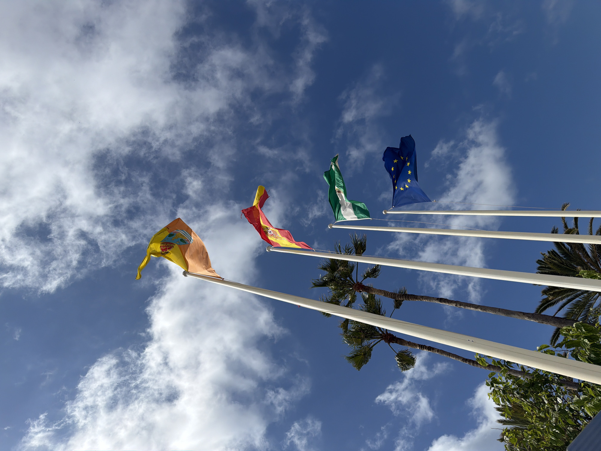 The image shows four flags on white flagpoles against a blue sky with scattered clouds. From left to right, the flags appear to be: a flag with a yellow and orange color scheme and a coat of arms; the Spanish flag (red, yellow, red); the Andalusian flag (green and white stripes); and the European Union flag (blue with yellow stars). Palm trees and other foliage are partially visible below the flagpoles. This scene is likely located in Arroyo de la Miel-Benalmádena Costa, Spain, given the presence of the Andalusian flag and typical Mediterranean vegetation.