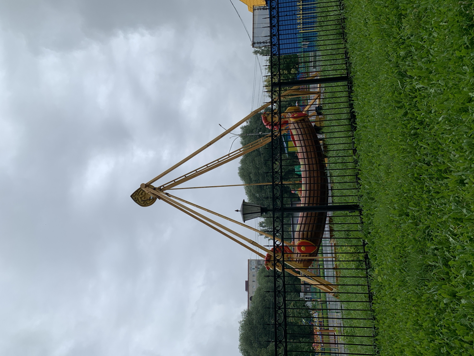 The image shows a ride in an amusement park in Prokopyevsk, Russia. The ride is a large, wooden boat suspended from a yellow frame. The boat is decorated with red and yellow shields and has what appear to be carved wooden figures on either side. The ride is surrounded by a black metal fence and lush green grass. In the background, there are trees, a building, and other park attractions. The sky is overcast and cloudy, suggesting it may be raining or about to rain. There are no people visible in the image.