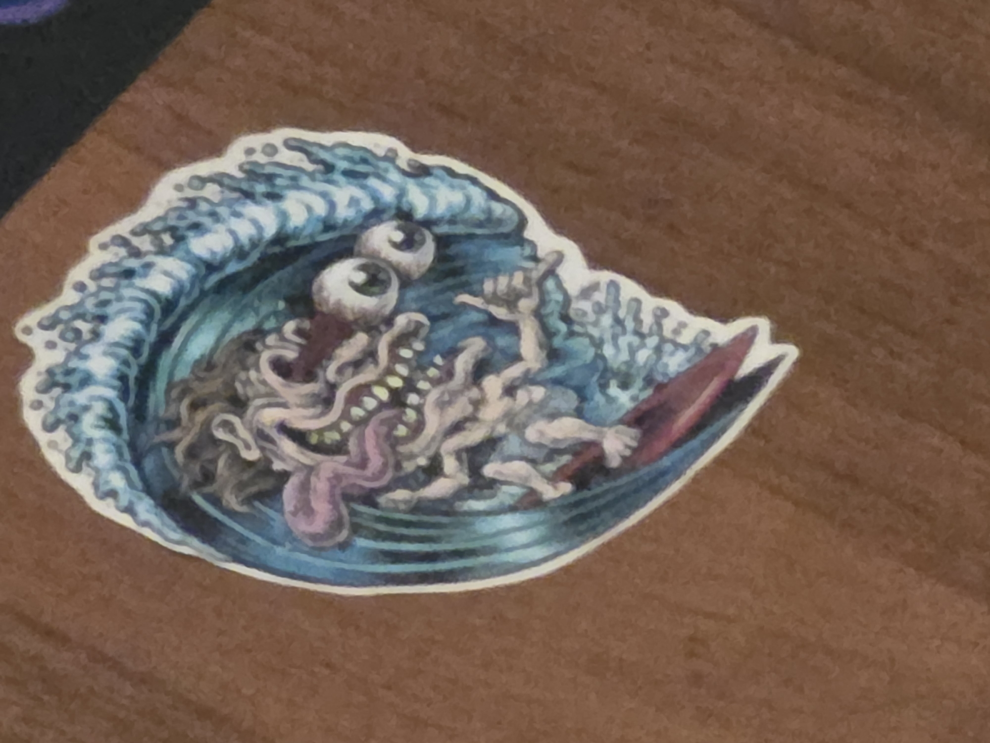 A sticker of a cartoonish creature surfing on a wave is stuck to a brown fabric surface. The creature has large, bulging eyes, a long, pink tongue sticking out, and a scraggly head of dark hair. Its body is thin and pale, with exaggerated limbs. It is riding a red surfboard through a stylized blue wave. The background of the sticker is white and jagged, giving the impression of a wave's crest. The fabric surface appears to be a textured cloth, possibly a bag or a piece of clothing.