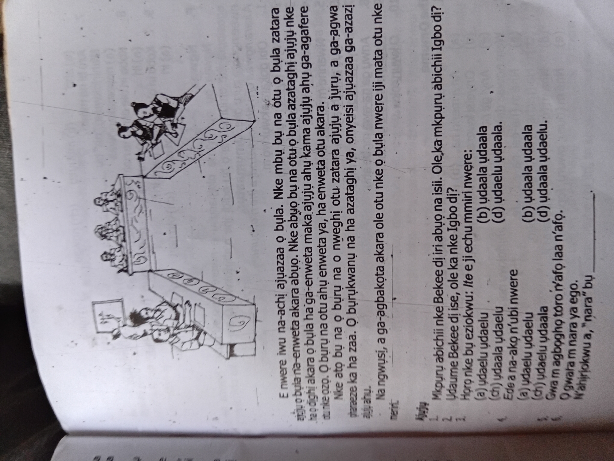 The image is a close-up, top-down view of an open textbook page featuring a hand-drawn illustration and text in an African language. The illustration depicts a formal gathering, possibly a court or council meeting, with several individuals seated at tables or desks. One group of figures is seated on a raised platform or behind a table, facing another group seated at a long, curved table. The environment appears to be an interior space, suggested by the desks and seated figures. The text is arranged in paragraphs and a numbered list of questions. The language appears to be Igbo, as indicated by phrases like "Nkpor, Nigeria" in the prompt. The specific location of Nkpor, Nigeria, is not visually represented in the image itself but is provided contextually. The overall impression is of an educational material, likely a textbook for learning the Igbo language, with the illustration serving as a visual aid for the accompanying text.