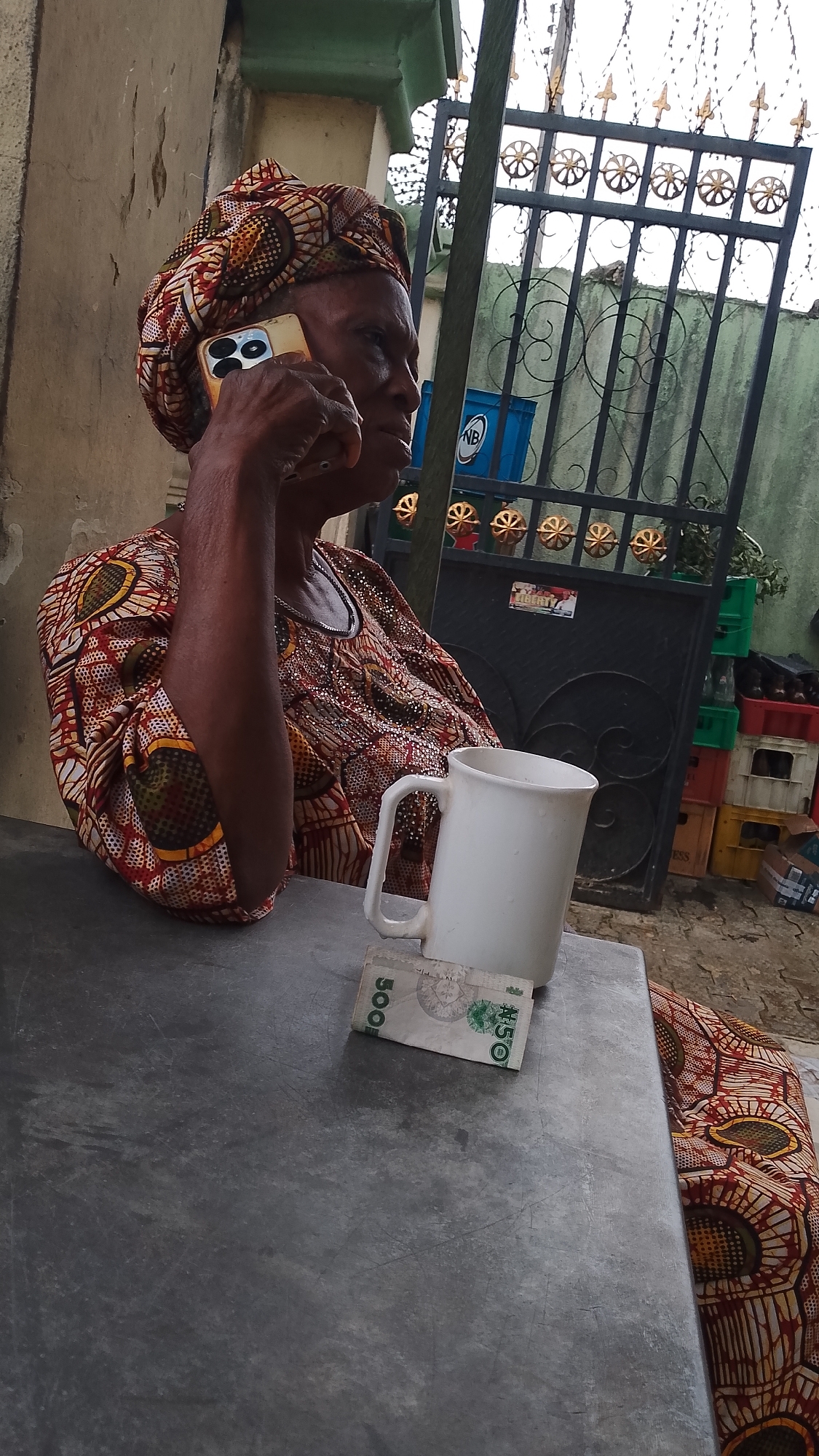 An older woman is seated at a gray table in Asubi, Nigeria, talking on a cell phone. She is wearing a patterned headwrap and a matching top. A white mug and two bills, one for 500 and the other for 50, are on the table beside her. In the background is a gate with decorative elements, and stacked crates.