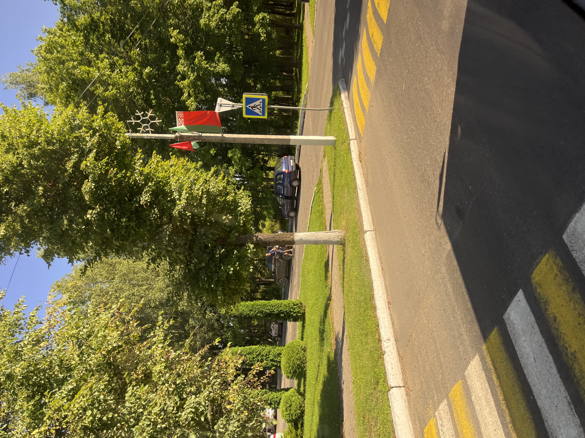 The image depicts a street scene in Myadzyel, Belarus, on a sunny day. In the foreground, a pedestrian crossing with yellow and white stripes is visible on the asphalt road, casting a shadow. To the left of the crossing, a grassy area with a curb separates the road from a sidewalk and a landscaped section with manicured bushes and tall, conical evergreens. A tree with a white-painted lower trunk stands near the sidewalk.
Further back, several cars are parked or driving. A blue sedan with its door open is parked on the right side of the road. A darker colored SUV is also visible, with a person standing next to it. In the background, more trees form a dense line, indicating a park or wooded area.
A utility pole stands prominently on the right side of the image, adorned with flags and a decorative snowflake-like ornament. Two flags are visible, one red and green, and another red flag. A pedestrian crossing sign, a yellow square with a blue border and a white silhouette of a person walking, is mounted on a pole next to the utility pole. The sky above is clear and blue. The overall impression is a well-maintained urban environment on a bright summer day.