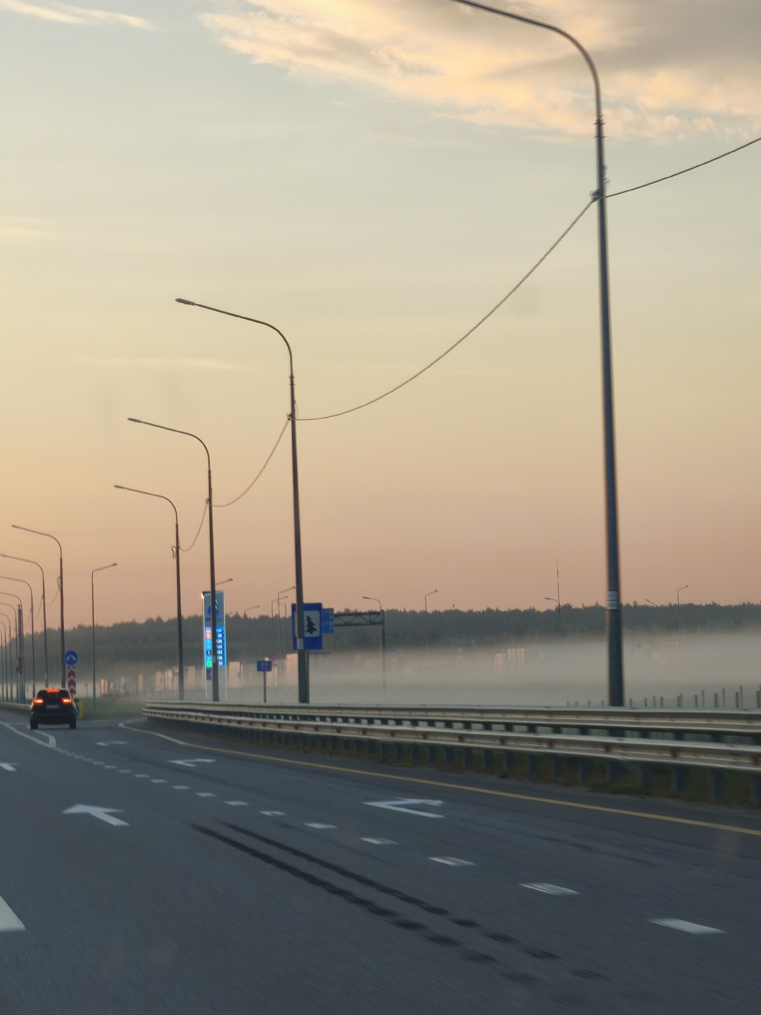 The image shows a highway at dawn in Bolshiye Dvory, Russia. A single car is driving on the road, and there are several streetlights lining the highway. The sky is a pale orange and yellow, with some clouds. There is fog in the distance, obscuring the treeline. Road markings include white arrows indicating lane direction and dashed white lines separating lanes. A guardrail runs along the side of the highway. Several road signs are visible, including a blue sign with white text and a blue sign with a white tree symbol. The overall scene suggests a quiet, early morning on a major road.