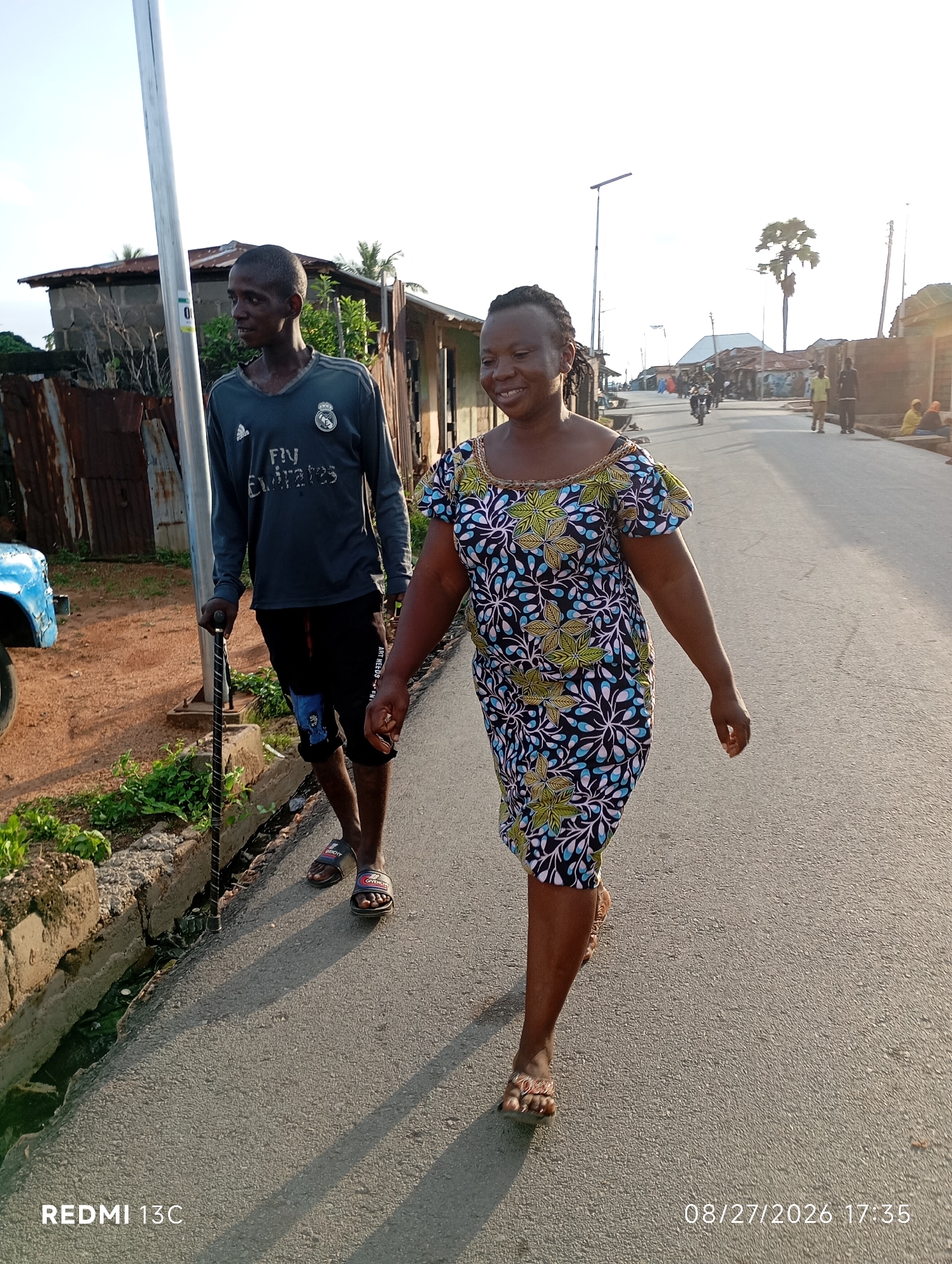 The image depicts an outdoor scene in the city of Shendam, Nigeria. In the foreground, two individuals are walking on a paved road. On the left, a young man, appearing to be of African descent, is walking with a cane, wearing a long-sleeved grey t-shirt with "FLY EMIRATES" printed on it and black shorts. He has a slender build and short hair. To his right, a woman, also appearing to be of African descent, is walking in a colorful patterned dress. She has a warm smile and her hair is styled in braids. The background shows a street lined with buildings and a few trees. The sky is bright and appears to be either early morning or late afternoon. The overall impression is a casual moment captured on a typical street in Shendam.