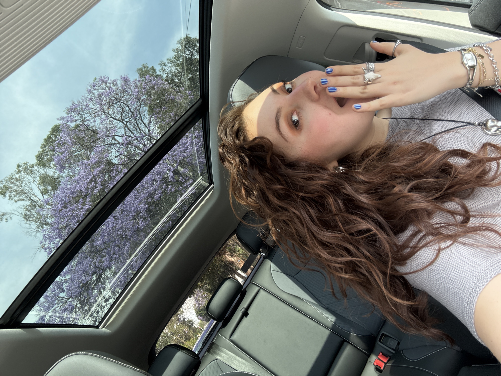 The image depicts a young woman inside a car with a sunroof, likely in Mexico City, Mexico, as indicated by the Jacaranda trees visible through the sunroof. She has brown, curly hair, fair skin, and wears a light grey top. Her nails are painted with blue stripes. She wears several rings on her right hand and a watch and bracelets on her right wrist. Her mouth is slightly open and her hand is raised to her mouth, suggesting surprise or shock.
Through the sunroof, a blossoming Jacaranda tree with violet-purple flowers is visible against a partly cloudy sky. The interior of the car features black leather seats, a visible seatbelt, and grey interior trim. The image is taken at an angle, and the camera appears to be held above her.