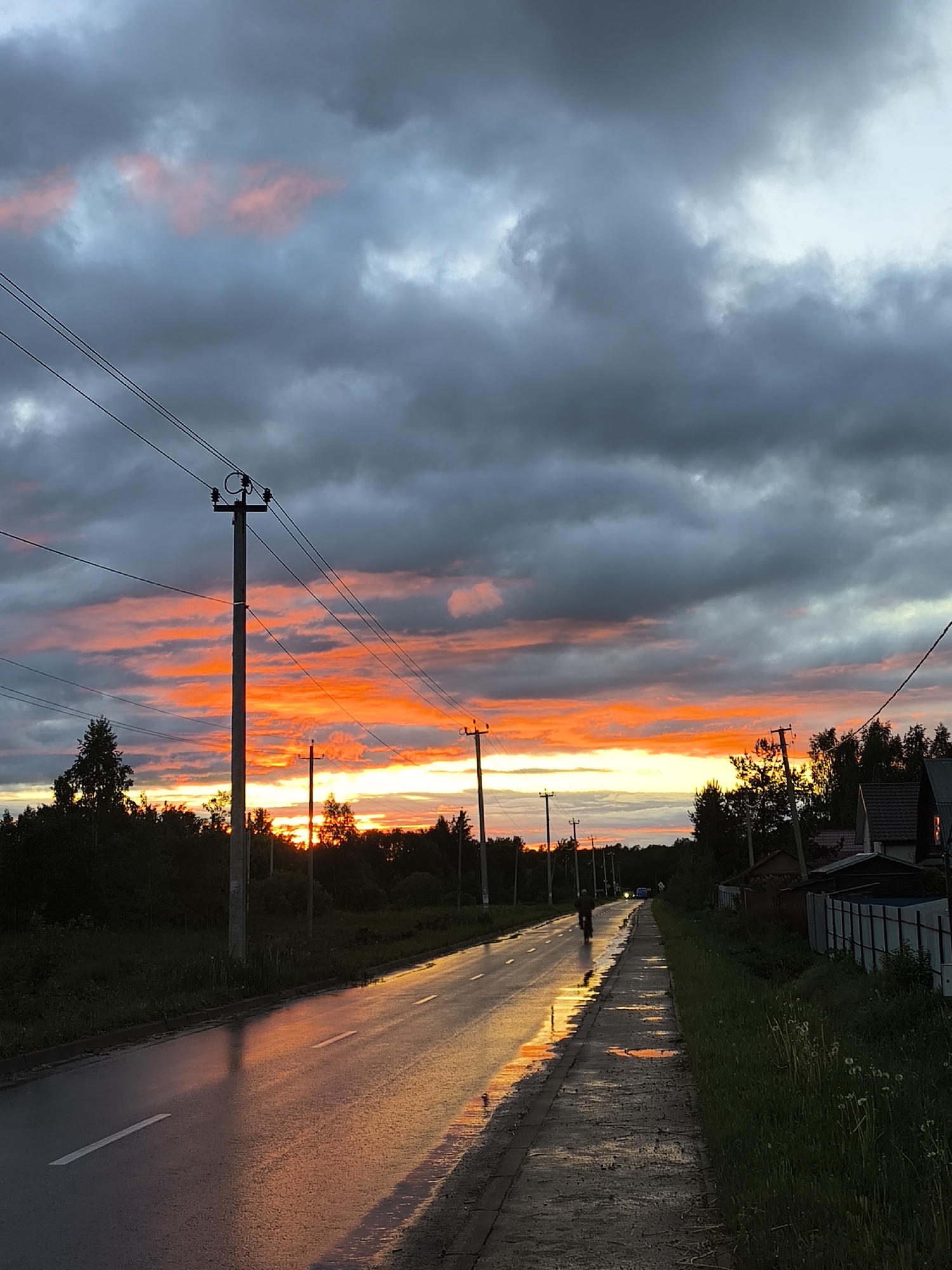 The image shows a road reflecting the vibrant colors of a sunset. The sky is a mix of dark, heavy clouds and streaks of orange and pink light. Power lines and poles run along the left side of the road. The road itself is wet, reflecting the colors of the sky. A sidewalk runs alongside the road on the right, with patches of standing water and greenery. A cyclist can be seen in the distance on the road. On the right side of the image, there is a fence and a house partially visible behind it. The image is taken at dusk. The location is a road with trees and houses typical of a rural or suburban area.
