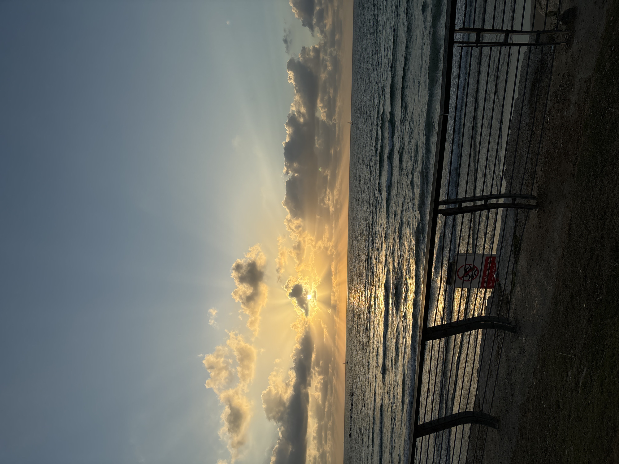 This image depicts a serene sunset over the Mediterranean Sea in Tel Aviv, Israel. The sun, partially obscured by clouds, casts golden rays across the water, creating a shimmering reflection. A sailboat can be seen in the distance, adding a touch of tranquility to the scene. In the foreground, a metal railing borders a sandy embankment, suggesting a promenade or walkway. A small sign with a prohibition symbol is attached to the railing. The sky is a gradient of blue and orange, with scattered clouds adding texture and depth. The overall atmosphere is peaceful and contemplative, evoking a sense of calm and natural beauty.