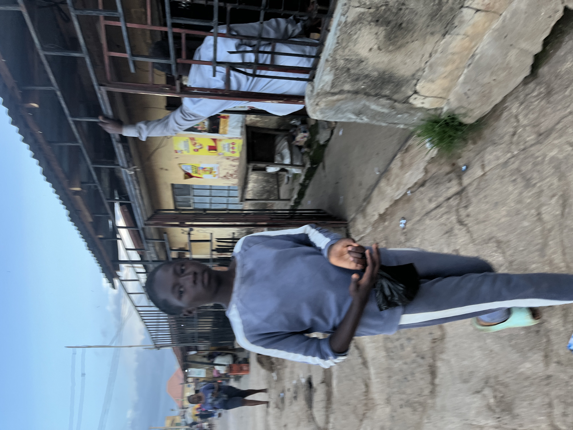 A young person, appearing to be of African descent, walks towards the camera on a paved path. They are wearing a blue long-sleeved shirt with white stripes on the sleeves and matching blue pants with a white stripe down the side of each leg. They are holding a black plastic bag in their right hand and have their left hand open. They are wearing light blue sandals.
In the background, a man in a light-colored shirt is visible inside a structure with a corrugated metal roof and metal bars. He is reaching up with his right arm. Behind him, the wall of the building is yellow and has posters on it. To the left of the young person, another person is walking away from the camera. The environment appears to be an urban setting in Jos, Nigeria, with a slightly overcast sky. The ground is concrete and there is some debris scattered on it.