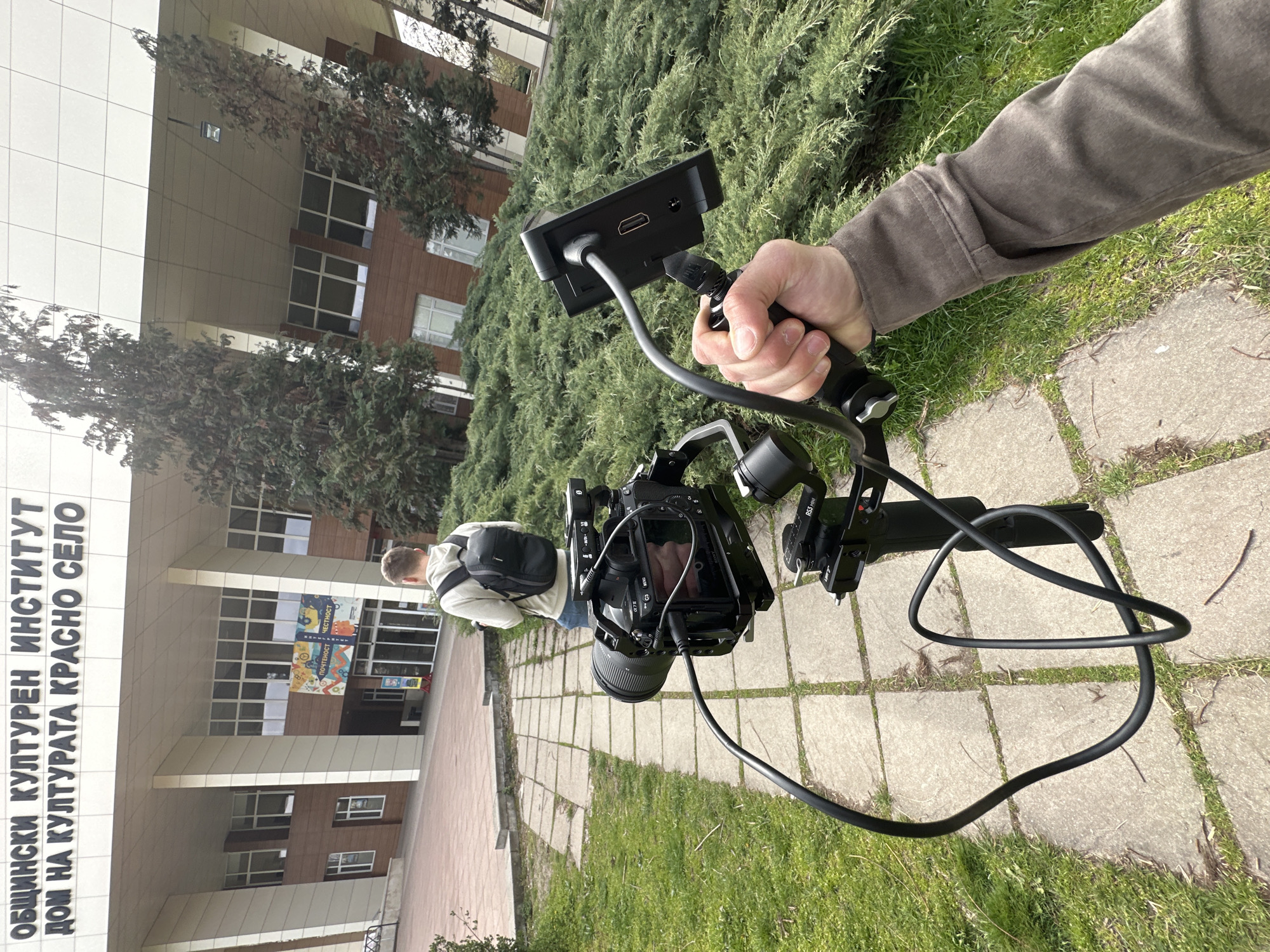 The image depicts a person holding a camera with an attached monitor, likely a video recording setup, in an outdoor environment. The camera is mounted on a gimbal stabilizer. A black cable connects the camera and monitor. In the background, there's a building with a sign in Cyrillic script; part of the sign reads "ОБЩИНСКИ КУЛТУРЕН ИНСТИТУТ ДОМ НА КУЛТУРАТА КРАСНО СЕЛО," which translates to "Municipal Cultural Institute House of Culture Krasno Selo." A person with a backpack is walking in front of the building. There are trees and grass around the area. The location is likely Krasno Selo district in Sofia, Bulgaria, based on the building signage.