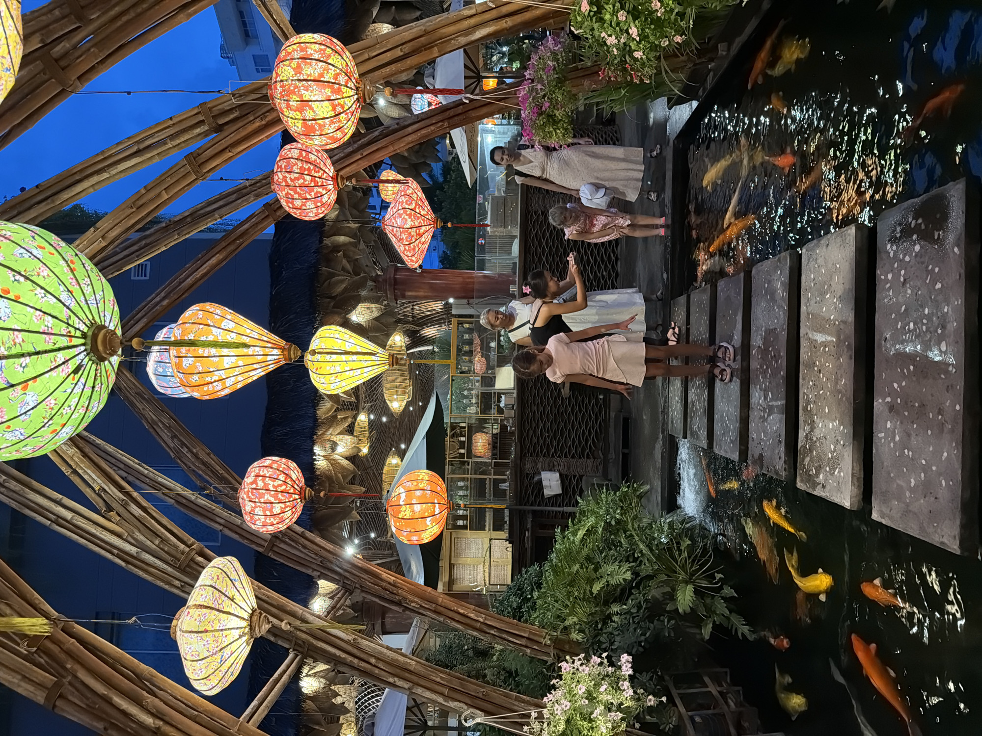 In Nha Trang, Vietnam, a vibrant scene unfolds under a canopy of traditional Vietnamese lanterns, illuminating a tranquil koi pond. The pond, teeming with a variety of colorful koi fish, is crossed by a path of large, flat stepping stones. On the path, two young girls, one in a pink dress and the other in a floral dress, are observed. An adult woman in a long, cream-colored dress stands nearby, holding a white cap, while another woman in a white dress is bent over, interacting with the younger girl. The background reveals a bar area with shelves stocked with bottles and glassware, suggesting a restaurant or entertainment venue. Lush green foliage and flowering plants add to the tropical ambiance. The architecture features natural bamboo structures that support the decorative lanterns, creating a festive and inviting atmosphere.