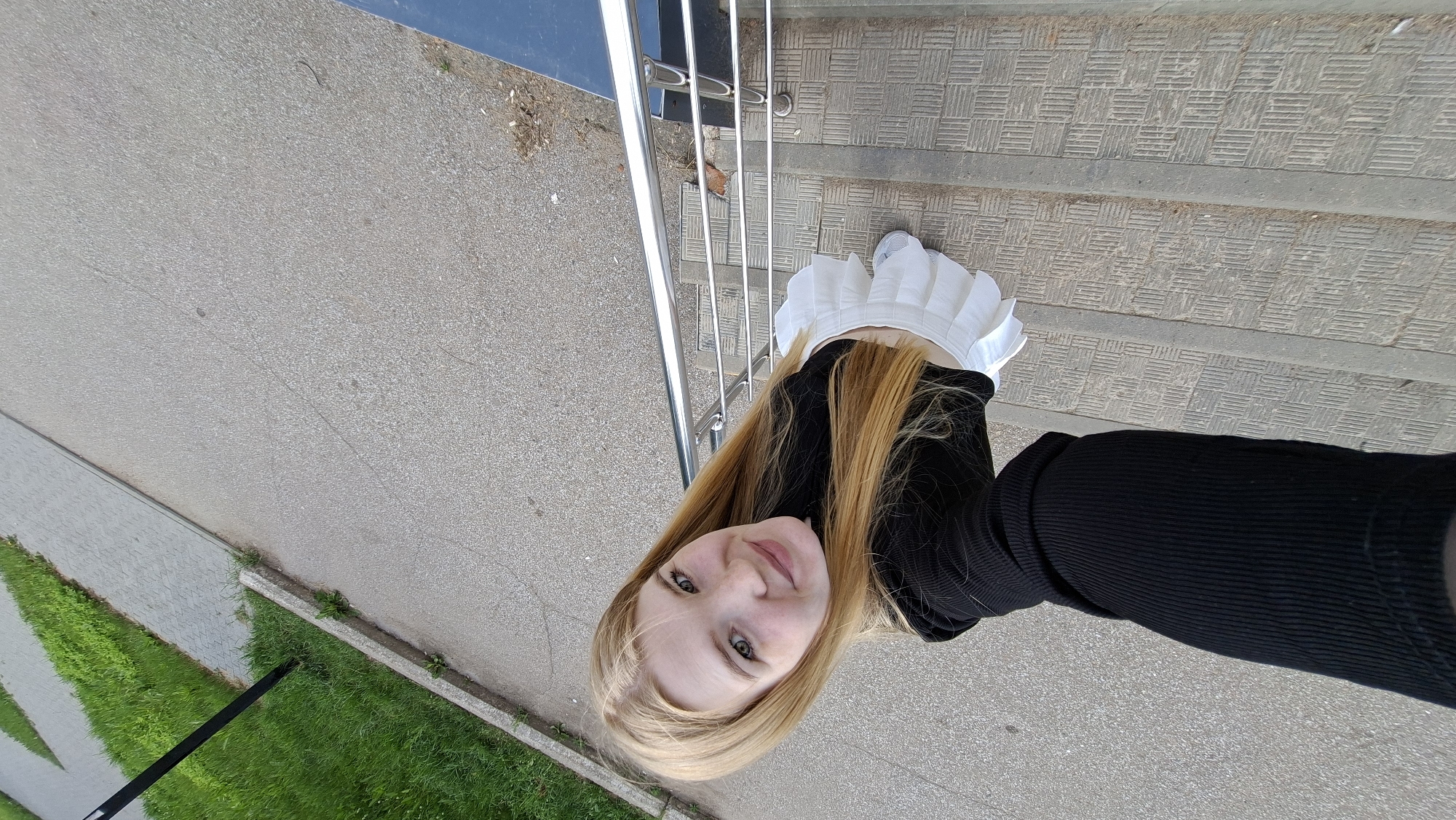 A young blonde woman with fair skin and green eyes is taking a selfie outdoors. She is wearing a black ribbed long-sleeved top and a short white pleated skirt. Her hair is long and blonde. She is standing on a textured grey outdoor staircase with a silver railing. To the left of the stairs, there is a paved pathway and a strip of green grass. The scene appears to be in a public outdoor space. The woman is looking up at the camera and smiling. The image is taken from a high angle, looking down at the woman.