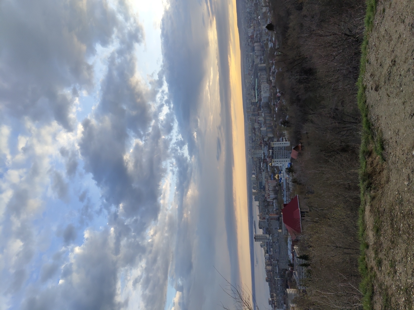 The image shows an elevated view of Saratov, Russia, looking towards the Volga River under a cloudy sky. The city sprawls along the riverbank with a mix of high-rise and low-rise buildings. A church with golden domes is visible amidst the urban landscape. In the foreground, there is a dirt path and vegetation on the edge of the hill from which the photo was taken. The lighting suggests either sunrise or sunset.