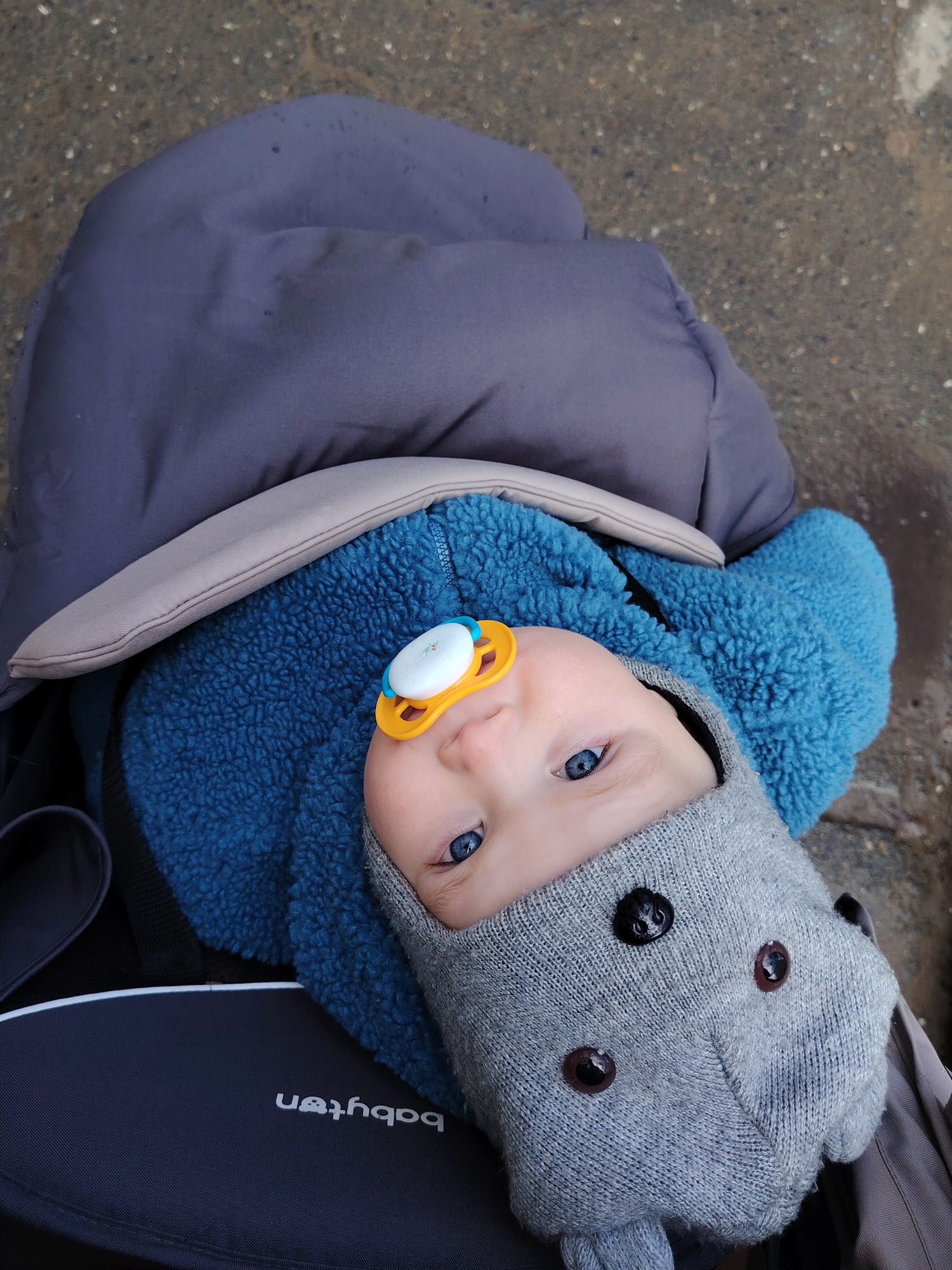 A baby lies in a dark-colored stroller with the brand name "babyten" visible, possibly in the city of Chaikovsky, Russia. The baby wears a light gray knitted hat with button eyes, a blue fleece jumpsuit, and has a pacifier in their mouth. A gray-blue blanket partially covers the baby. The background appears to be a wet or damp surface, perhaps a sidewalk or pavement. The stroller is oriented towards the camera.