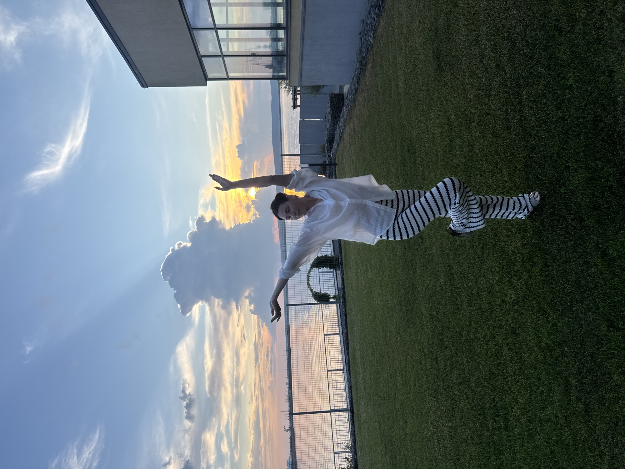 A woman stands on one leg in a yoga pose in a grassy courtyard. She is wearing a white button-down shirt and black and white striped pants. Her arms are raised, with her left arm extended upwards and her right arm bent at the elbow. She is looking to her left. Behind her, a body of water reflects the sunset sky, which is filled with dramatic clouds. A fence separates the courtyard from the water. To the right of the woman is a modern building with large windows. In the distance, industrial structures are visible along the shoreline. The scene appears to be set in Chaikovsky, Russia, likely near the Kama River.
