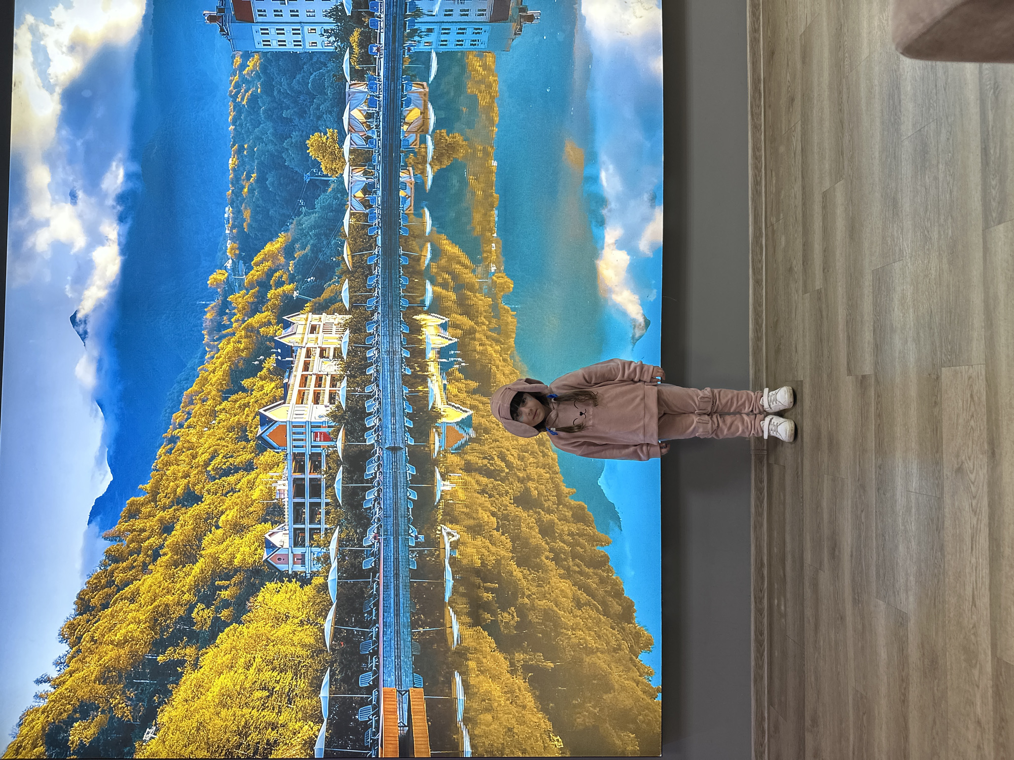 A young girl in a pink hooded jumpsuit and white sneakers stands in front of a large screen displaying an inverted image. The image shows a mountainous landscape with autumn foliage and buildings, likely a reflection in a body of water. The scene is suggestive of a location in Sochi, Russia, given its known geography. The girl is looking towards the camera, while the screen behind her dominates the visual space. The background includes a grey wall and a wooden floor.