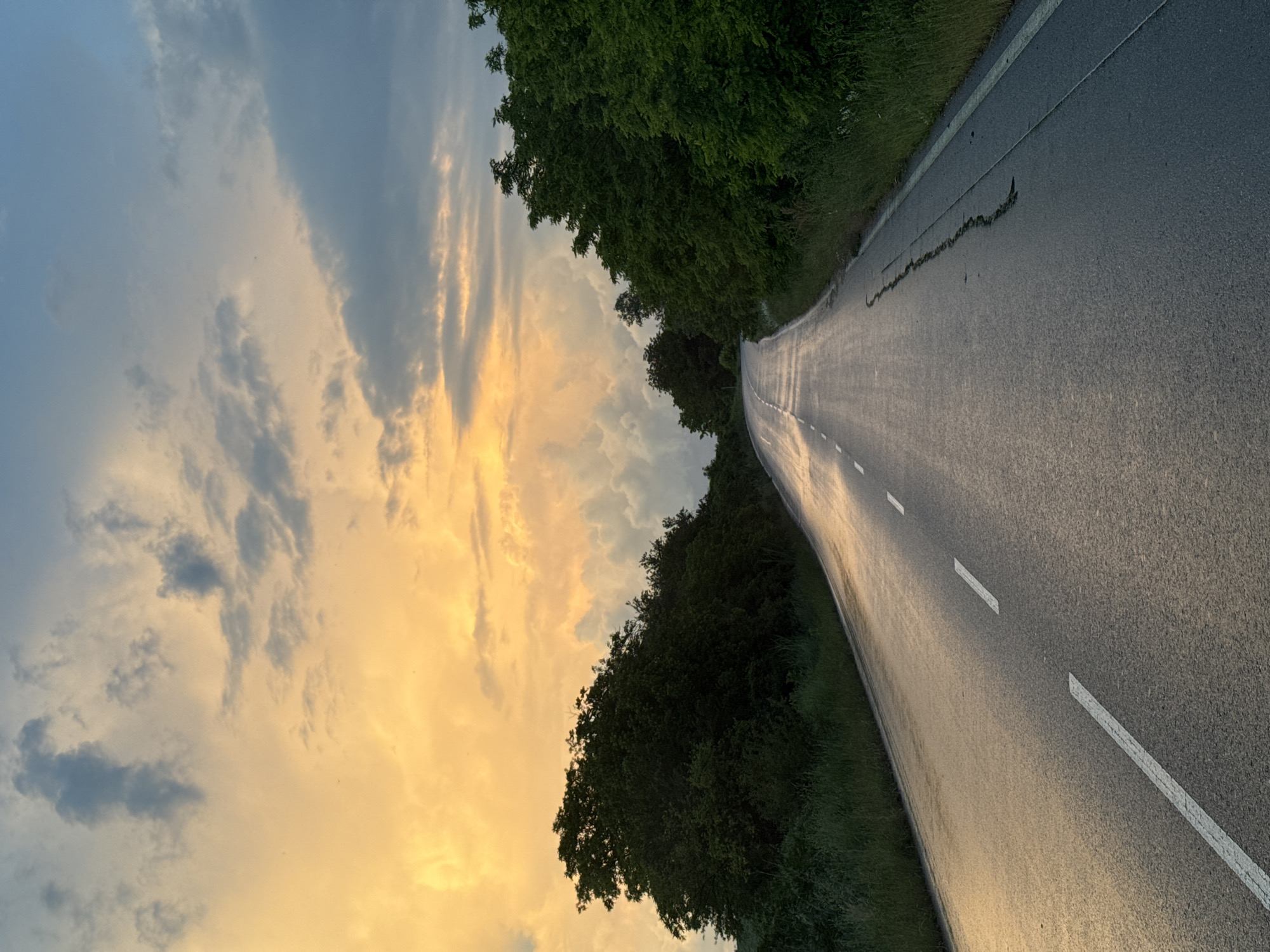 The image shows a road stretching into the distance under a sky filled with clouds illuminated by the setting sun. On either side of the road are trees and vegetation. The road has white lane markings. The photograph is rotated; the sky appears to be on the left side of the frame, and the road runs roughly from the top to the bottom. The specific location is not identifiable as Strazhitsa, Bulgaria, solely from visual information in the image.