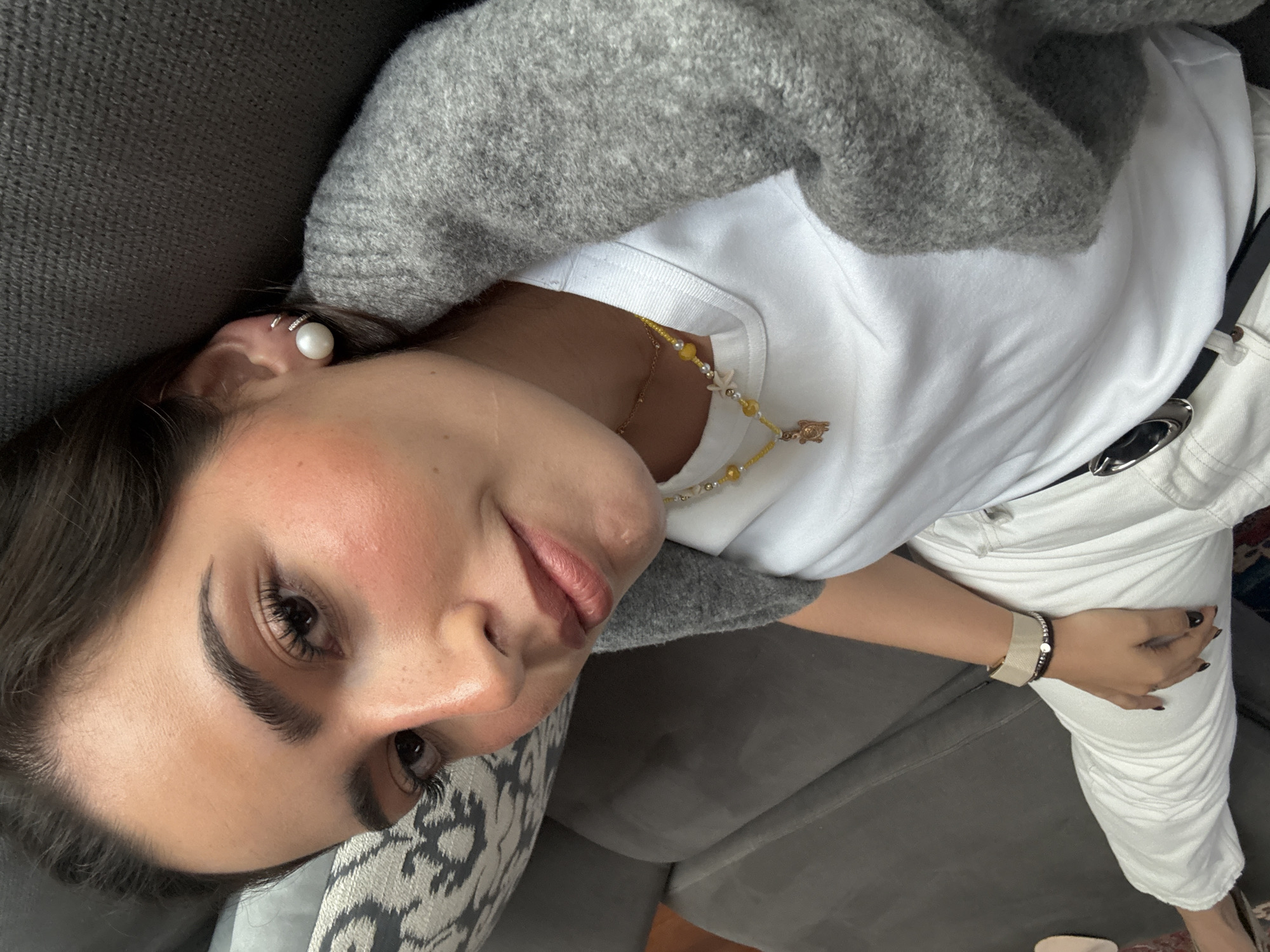 The image is a close-up, selfie-style shot of a woman lying on a grey sofa. She is tilted towards the camera. She has dark hair and is wearing a pearl earring in her left ear. She is wearing a white t-shirt, a grey cardigan, and white pants with a black belt. A beaded necklace with yellow and white beads is visible around her neck. She is wearing a beige and black bracelet on her left wrist. Her fingernails are painted black. The location in San Bernardino Tlaxcalancingo, Mexico, cannot be determined from the visual information provided.