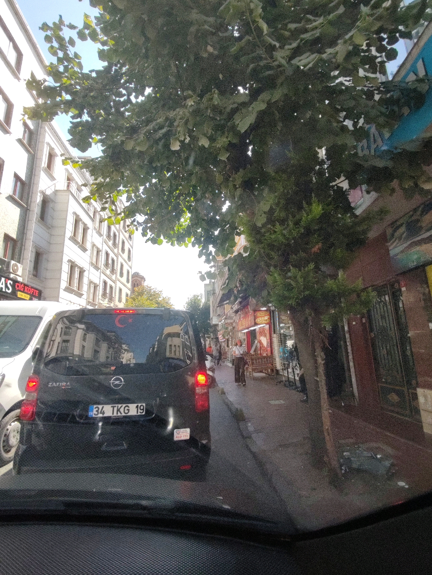 The image shows a street scene in Istanbul, Turkey, likely in a busy urban area with buildings lining the street. In the foreground, the rear of a black Opel Zafira car is visible, with the license plate "34 TKG 19" clearly displayed. A Turkish flag, consisting of a red background with a white star and crescent moon, is affixed to the rear window.
To the left of the car, part of a white vehicle is visible. Beyond the cars, buildings with multiple stories and windows are present. Trees with green foliage overhang the street, partially obscuring the view of the sky, which appears to be blue.
On the right side of the street, there are shops with their entrances and displays visible. One shop has a prominent sign in Turkish, and another appears to be selling goods. A few pedestrians are walking on the sidewalk. The street itself appears to be paved, and there are signs of urban activity. The overall impression is a snapshot of daily life in a bustling Turkish city.