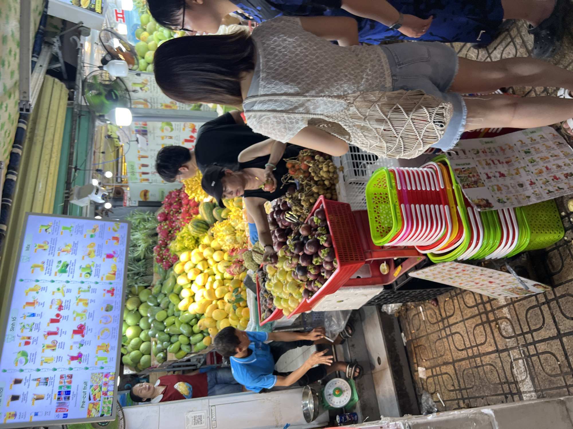 The image depicts a vibrant fruit stand in Nha Trang, Vietnam. In the foreground, a man in a blue shirt is weighing fruit, with a digital scale to his left. Behind him, a woman in a black tank top is arranging mangosteens in a red basket, holding a small red fruit in her hand. Various other fruits, including rambutans and what appear to be dragon fruits and pineapples, are also displayed in baskets and piles. A large digital menu board hangs above the fruit stall, displaying images and text in Vietnamese, listing various fruit juices and mixes. The background shows more fruit displays and a lively market atmosphere, with awnings overhead. The ground is tiled, and there are stacks of colorful plastic baskets to the right. Two women are visible in the mid-ground, one in a white lace top carrying a woven bag, and another in a blue patterned shirt. The overall scene is one of a busy, colorful, and bustling outdoor market.