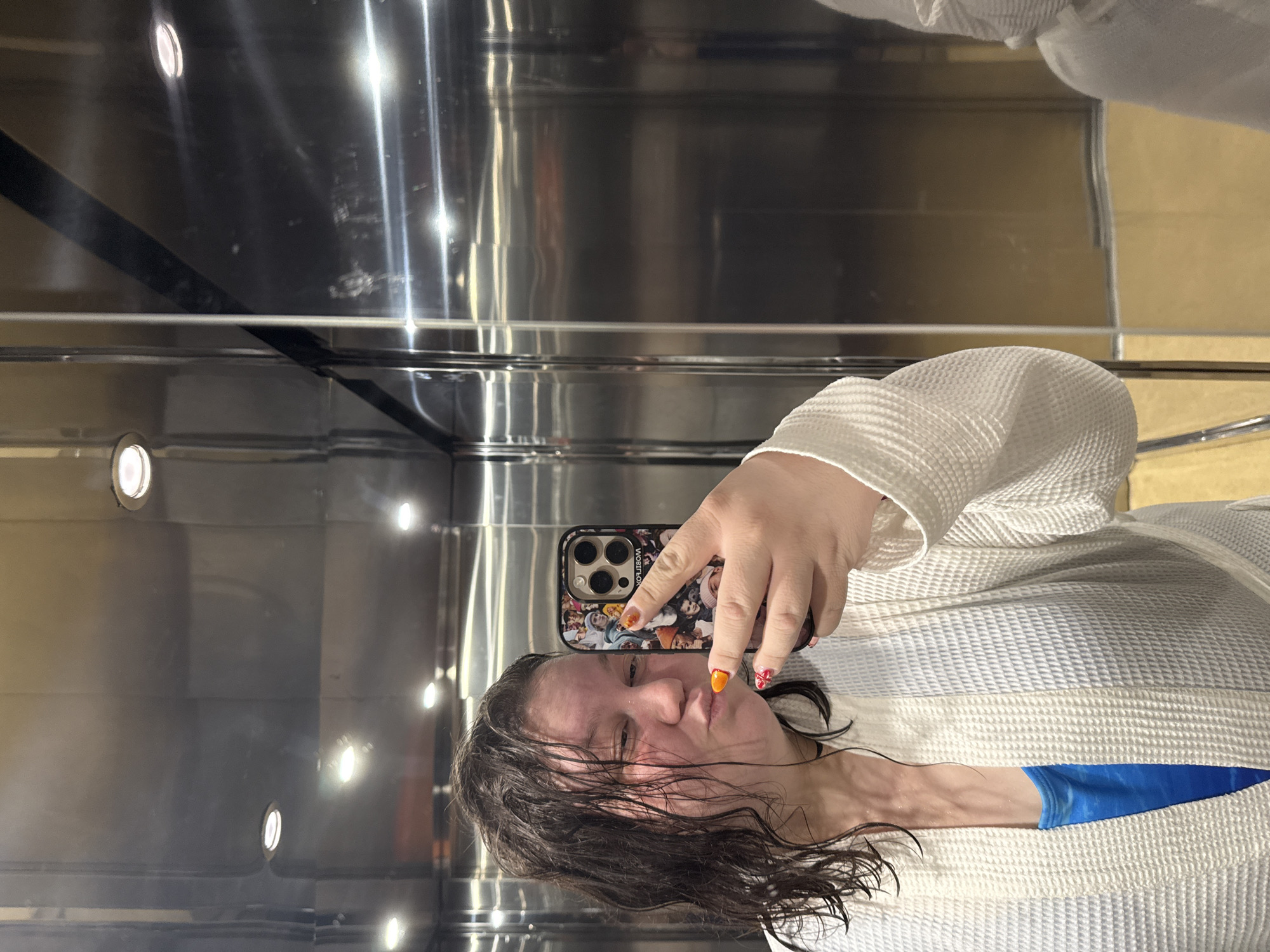 The image, captured in Tulovo, Bulgaria, depicts a female subject taking a mirror selfie within what appears to be an elevator.
The subject is a woman with dark, wet, and visibly disheveled hair, which hangs loosely around her face and shoulders. Her skin appears somewhat flushed. She is making a puckering or "kissing" expression with her lips and her eyes are directed towards the smartphone screen. She is wearing a white, waffle-knit bathrobe, partially open to reveal a blue garment underneath, possibly a swimsuit top or tank top. Her left hand is prominently visible, holding a dark-colored smartphone, likely an iPhone model due to the distinct triple-lens camera array. The phone is encased in a protective cover featuring a collage of multiple small photographic images, predominantly depicting various faces and individuals. The word "WOMBLOOK" is discernibly printed in reverse on the phone case, indicating a reflection. Her visible fingernails are painted; the thumbnail is a solid bright orange, while the index finger displays a red and white patterned design.
The environment consists of highly reflective, brushed metallic surfaces forming the walls and ceiling, consistent with an elevator interior. Several circular, recessed downlights are visible on the ceiling, emitting bright light. The metallic surfaces show various reflections, including horizontal light streaks and apparent smudges or marks. A vertical dark seam or panel division is visible on the right side of the reflective surface. The lower portion of the reflection on the right wall indicates a muted, light brown or beige color, potentially a floor or lower wall panel outside the direct frame.