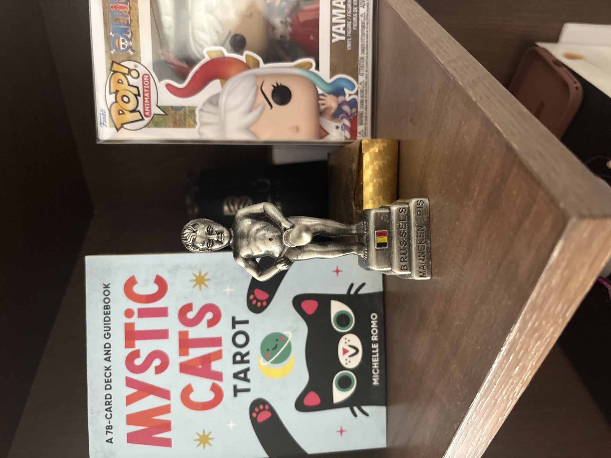 The image shows a shelf with a "Mystic Cats Tarot" deck and guidebook, a Manneken Pis statuette labeled "Brussels," and a Funko Pop! figurine in its box. The shelf is made of wood and appears to be part of a larger shelving unit.