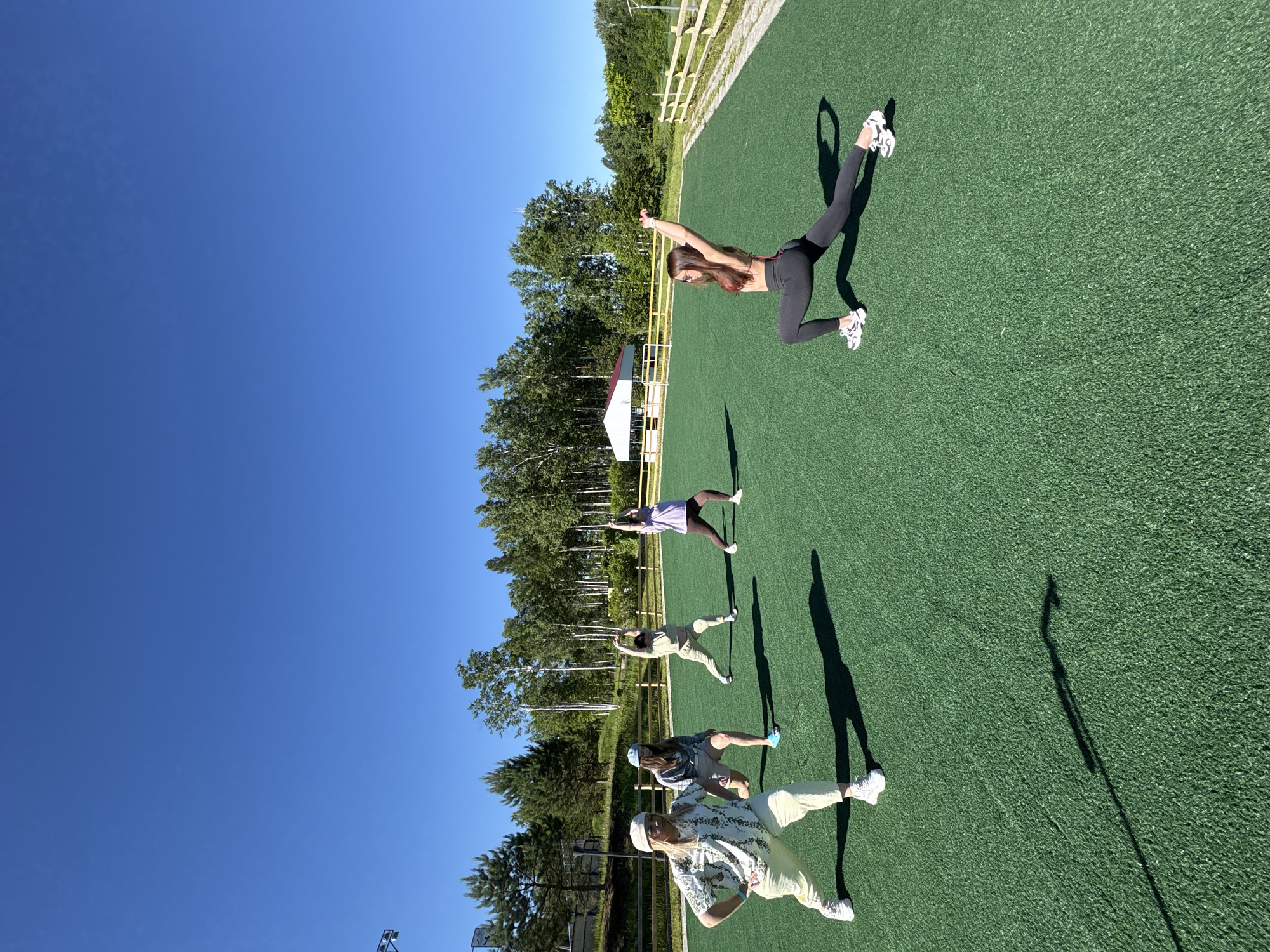 Five individuals are engaged in a physical activity session outdoors on a bright, sunny day. The setting appears to be a sports field with artificial turf, surrounded by trees and a wooden fence. In the background, a white building with a red roof is visible amongst the trees. The individuals are positioned in a semi-circle on the green field, performing what seem to be stretching or yoga poses. One person is in the foreground on the left, wearing a light-colored outfit and a hat, with their hands on their hips. To their right, another individual is in a lunge position. Further back, three more individuals are spread out, also performing similar poses. The sky above is clear and blue. The location is identified as Birobidzhan, Russia, suggesting an outdoor recreational area within or near the city.