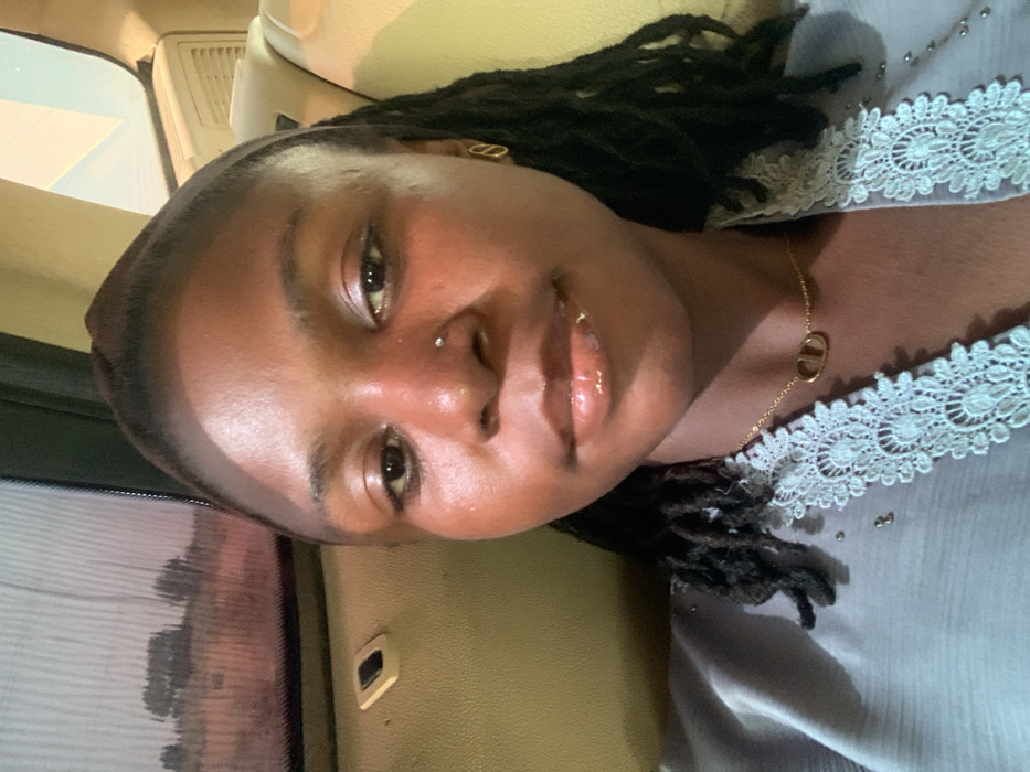 A close-up image depicts a young woman inside a vehicle, appearing to be in a selfie format. The woman has dark skin, dark dreadlocked hair pulled back, a small stud piercing on her right nostril, and a small labret piercing on her lower lip. Her lips are glossy. She wears a gold-colored necklace with a "CD" pendant and a gold-colored elongated oval earring in her visible left ear. Her attire is a light blue or gray garment featuring a white lace trim with a floral pattern and small metallic embellishments. The vehicle interior is tan/beige. A window on the left side shows a blurred exterior scene with dry, light-colored ground and sparse dark vegetation or trees, under what appears to be a bright sky. A beige panel with a black rectangular button or switch is visible below the window. The image does not contain specific visual identifiers to confirm the location as Gadam, Nigeria.