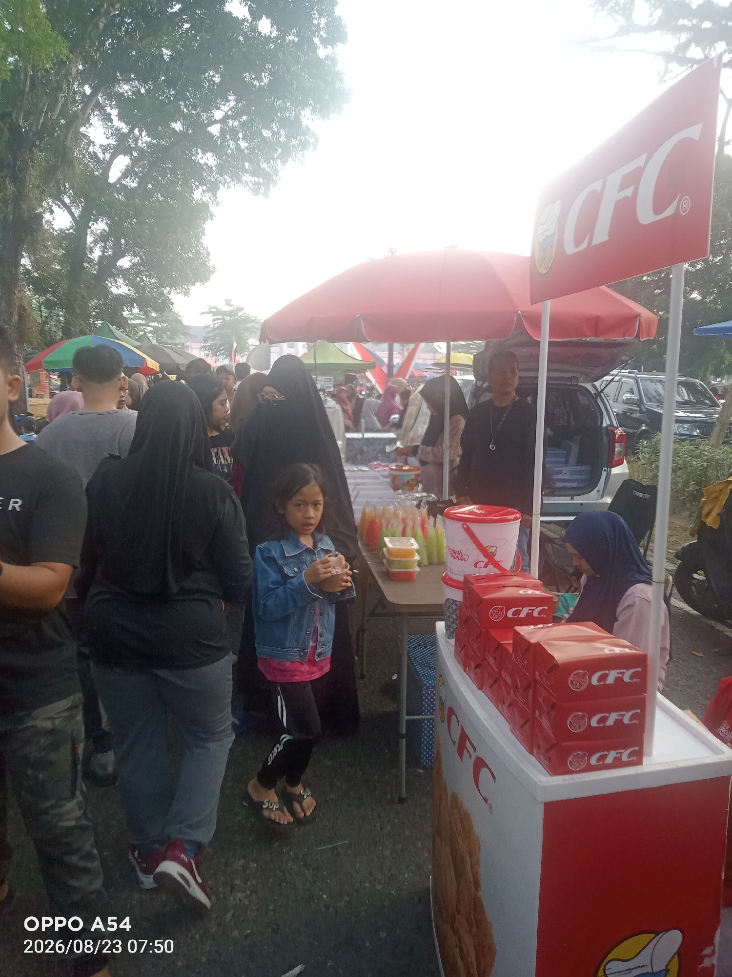 In Jambi City, Indonesia, a street scene unfolds under a bright sky, possibly late afternoon or early morning, with a hint of haze in the air. The foreground is dominated by a food stall displaying several stacks of red boxes, each bearing the "CFC" logo. Next to these boxes, a white cooler with "CFC" branding sits on a table. A young girl in a denim jacket is holding a cup, looking towards the camera. Adjacent to the food stall, a woman in a blue hijab is seated, appearing to assist with the sales.
Behind the stall, a man in a black shirt is seen standing, possibly a vendor. A red umbrella is open above part of the stall. Further back, other food stalls are visible with colorful umbrellas. People are milling about, some in traditional Islamic attire, suggesting a marketplace or a public gathering. Vehicles, including a dark SUV and a motorcycle, are present in the background, indicating a semi-urban or suburban setting. The overall atmosphere suggests a bustling outdoor market or a local event.