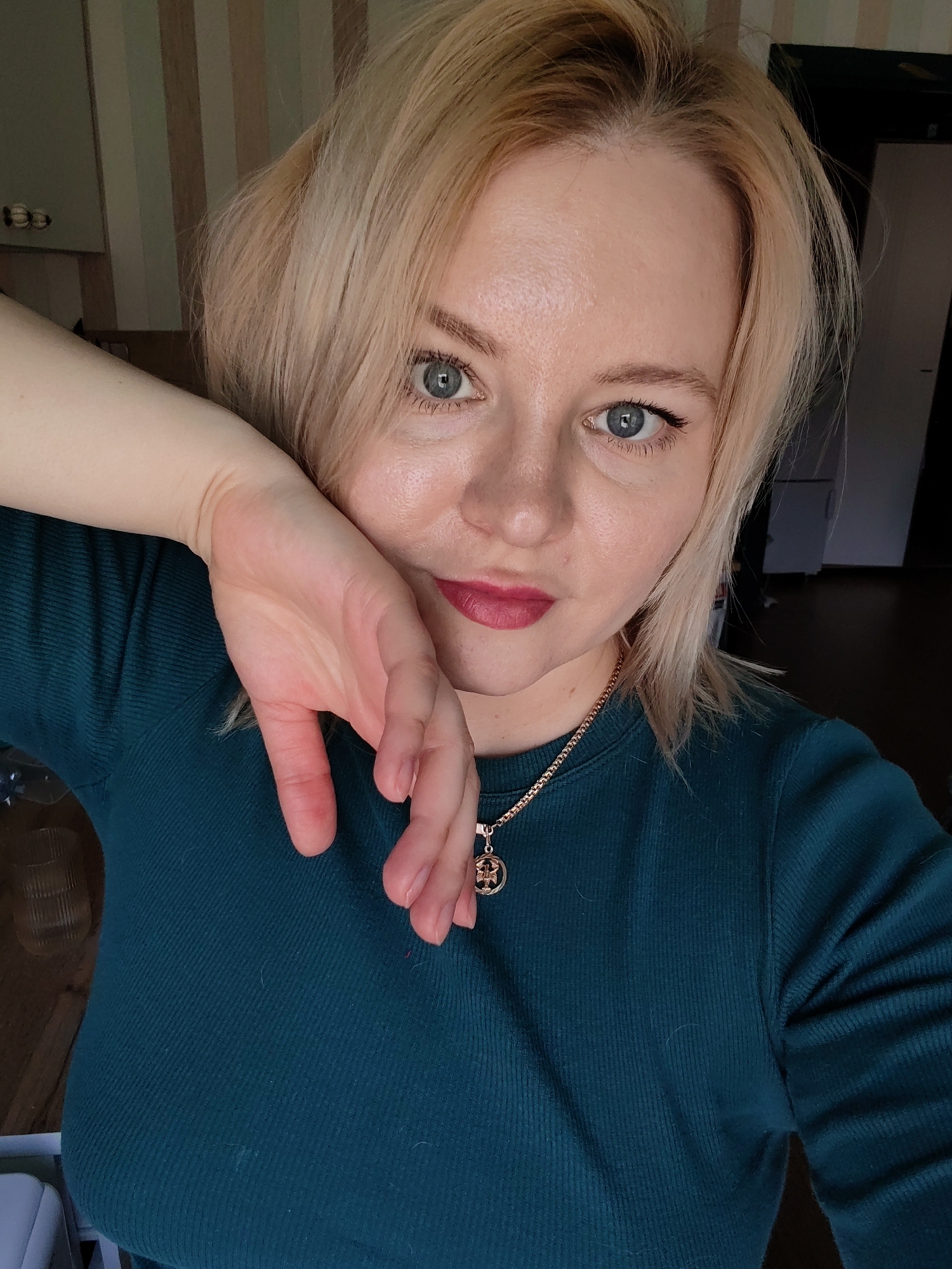 A blonde woman, appearing to be in her late 20s to early 40s, is captured in a close-up selfie taken indoors, likely in Izhevsk, Russia. She has short, slightly disheveled blonde hair, light blue or grey eyes with visible eyeliner and lashes, and fair skin. Her expression is neutral and direct as she looks into the camera. She wears a dark teal or green ribbed long-sleeved top and a gold chain necklace with a small, intricate circular pendant. Her left hand is raised, partially covering the lower left side of her face, with fingers slightly splayed, suggesting she is holding the camera for the selfie.
The indoor background reveals vertical striped wallpaper in shades of light green, beige, and white on the upper left. Below this, a dark cabinet or shelf unit is visible. On the right, a darker area suggests a doorway or large piece of furniture, with a dark wooden floor extending across the lower portion of the image. A light-colored object, possibly a piece of furniture, is partially visible in the lower left corner. The lighting is soft and even, indicating either natural daylight or well-diffused indoor lighting. There is no visible text in the image.