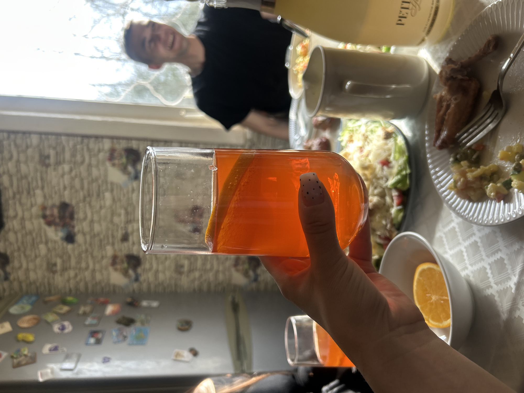 The image captures a casual indoor meal or gathering from a first-person perspective, with a hand holding a glass in the foreground. The scene is likely set in a dining area within a home in Шевлягино, Russia.
In the immediate foreground, a hand with light skin and white fingernails adorned with small black dots holds a tall, clear cylindrical glass filled with a vibrant orange beverage, possibly juice or a cocktail. An orange slice is visible inside the glass. Another similar empty glass is partially visible in the lower right corner.
A checkered white tablecloth covers a table laden with food and drinks. Visible dishes include a plate holding what appears to be a chicken leg or wing next to a portion of Olivier salad, and another plate with a green salad. A white bowl containing a half-slice of orange is also on the table. A white mug stands near the dishes. A bottle with a label showing "PETIT" (likely part of a sparkling wine or champagne brand like "PETIT PARIS") is also on the table, contributing to a celebratory or relaxed atmosphere.
In the background, a man with a smiling face, wearing a black t-shirt, is out of focus, suggesting he is seated at the table opposite the photographer. Bright natural light streams in from a large window behind him, indicating it is daytime. The walls are covered in a textured, light-colored wallpaper. To the left, a refrigerator is adorned with numerous magnets, a common sight in many homes.
The overall impression is one of a friendly and relaxed mealtime gathering. The bright lighting and smiling face suggest a pleasant and social occasion.