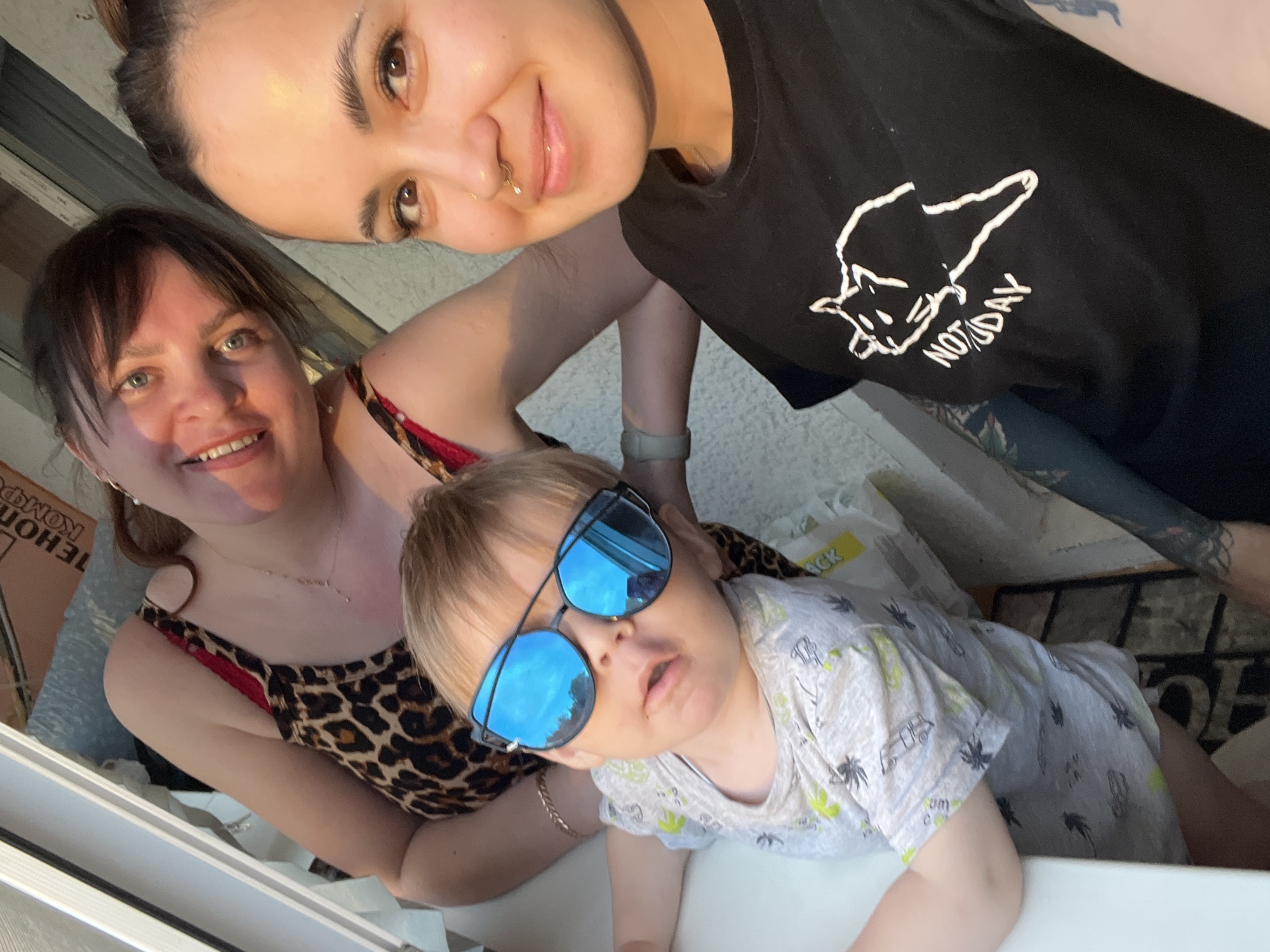 The image shows three people, two adults and one child, in what appears to be an indoor or semi-outdoor setting, possibly a balcony or by a window. The lighting suggests daytime.
On the left, a woman with dark hair and bangs is smiling, wearing a leopard-print top with red accents. Next to her, to the right, is a young child wearing a light-colored t-shirt with a pattern and large blue mirrored sunglasses. The child is looking towards the camera. On the far right, another person, also a woman with dark hair pulled up, is smiling and facing the camera. She is wearing a black t-shirt with a white graphic of a cat and the words "NOT TODAY" printed on it. She has visible tattoos on her arm. The background is somewhat blurred, showing a wall and possibly some objects. Based on the provided information, this scene is located in the city of Noviy, Russia.