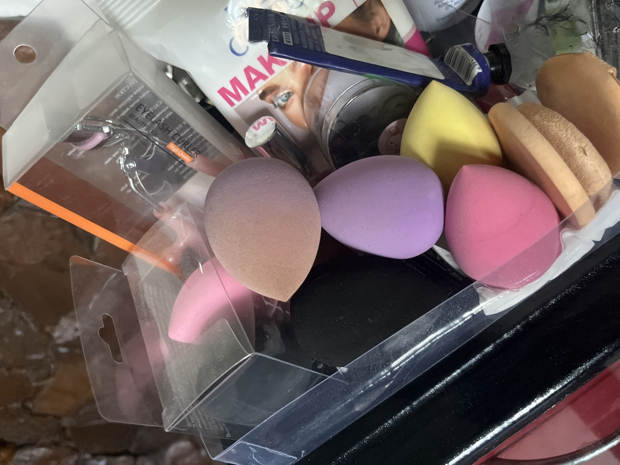 The image shows a close-up, overhead view of a disorganized collection of makeup sponges, an eyelash curler, and other beauty products piled together. The sponges are in various colors, including pink, purple, yellow, and a tan/orange hue. Some are still in their original clear plastic packaging. The eyelash curler is silver with pink accents and is housed in a clear plastic box. Other items visible include what appears to be a makeup palette with a person's eye on it, a tube or bottle with "MAKEUP" printed in large pink letters, and a blue tube with some text and a barcode. The background is a rough, textured surface that could be a wall. The lighting is somewhat dim and natural. There are no people visible in the image. The context suggests an indoor setting where makeup products are stored or kept. There is no information in the image to suggest the location is in Bohunove, Ukraine.