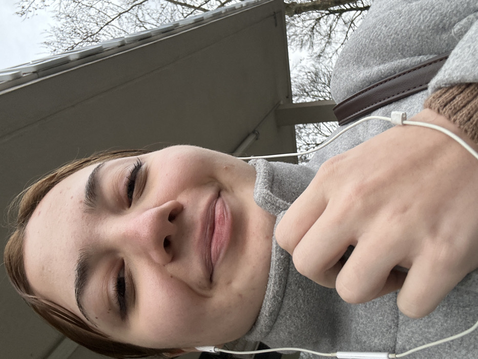 A low-angle selfie shows a person with fair skin, dark eyebrows, and closed eyes, suggesting a smile. They are wearing a gray turtleneck sweater and a gray coat with a brown strap. White earphones are visible, wrapping around the neck and chest. In the background, an unclear view of a building roof and bare tree branches against a cloudy sky can be seen.