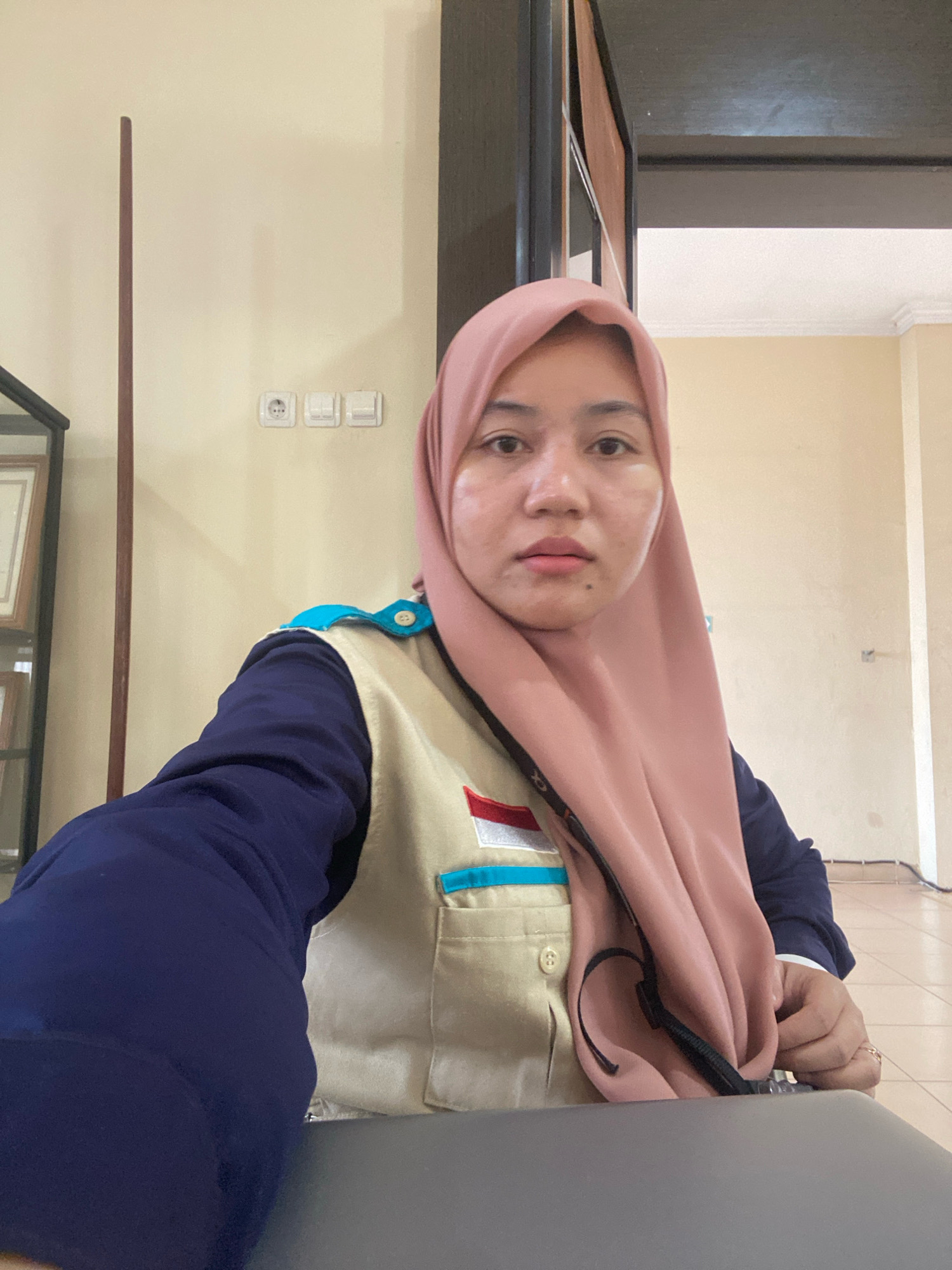 A young Indonesian woman wearing a pink hijab and a tan vest over a navy blue shirt is taking a selfie indoors. She has light brown eyes and light skin. The vest has a light blue collar and a pocket with a small Indonesian flag patch. She is seated at a grey table, with a dark brown wooden door frame visible behind her. The room has beige walls with electrical outlets and light switches. To her left, a tall, thin, dark brown pole stands near a glass display case with framed items. A black strap from a bag is draped over her shoulder and across her chest. Her right hand is visible, resting on her lap, wearing a ring on her index finger. The overall impression is an informal indoor portrait.