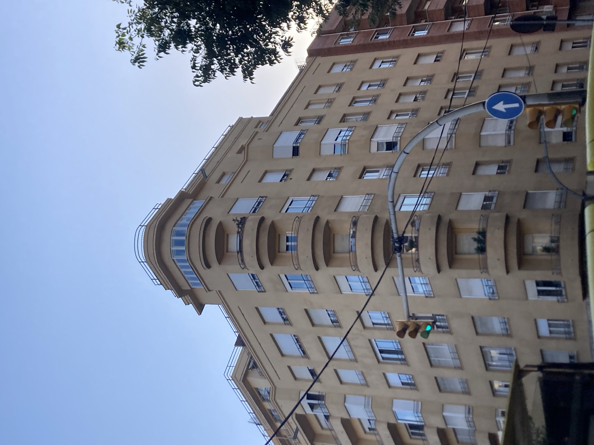 The image captures a street scene in Barcelona, Spain, focused on a light yellow, multi-story building with numerous windows and balconies. The architecture suggests an Art Deco or similar modernist style, characterized by curved balconies and a rounded corner on the upper floors. The building's facade is light yellow, and many windows have white blinds or shutters. A traffic light, showing a green light, is visible on the left side of the image, mounted on a curved metal pole. Below the traffic light, a blue circular road sign with a white arrow pointing upwards indicates a one-way street. The sky above is a clear, pale blue, with the upper branches of a dark green tree visible on the right side of the frame, partially obscuring the sky and the adjacent brick building. The overall lighting suggests it is either early morning or late afternoon, with a soft, diffused light. No people or vehicles are clearly discernible in the foreground or on the building itself.