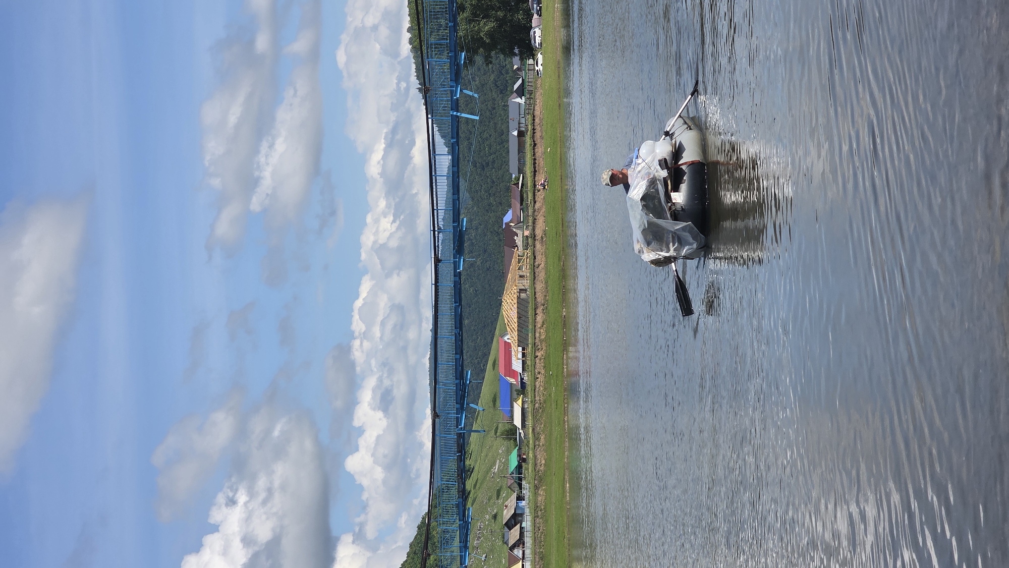 In the foreground, a person wearing a camouflage cap and a light-colored shirt is paddling a gray inflatable boat across a body of water. The boat is loaded with items covered by a translucent plastic sheet. The water's surface is rippled, reflecting the sky and clouds. In the middle ground, a village with houses of various colors, including red, blue, and green roofs, is situated on a grassy bank. A structure under construction with exposed wooden beams is also visible. A blue suspension bridge spans across a hill covered in greenery in the background, beyond which are forested mountains. The sky is a bright blue with scattered white clouds. The scene is located in the village of Tash-Asty, Russia.