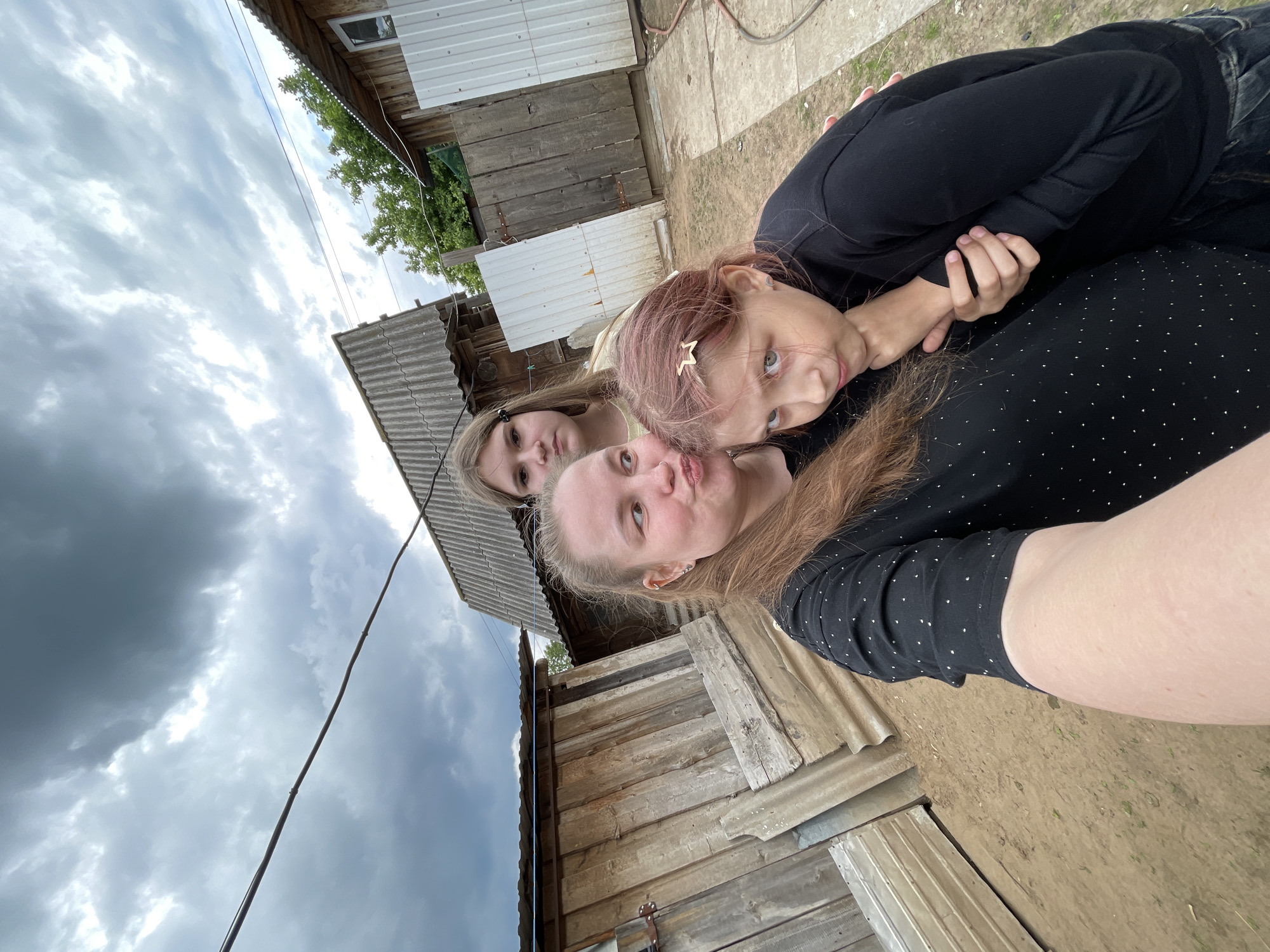A selfie photograph taken outdoors in Балдейка, Russia, features three individuals positioned closely together. A woman with red hair wearing a black, polka-dot shirt is in the foreground, making a kissing face toward the camera. Behind her to the left is a girl with blond hair, wearing dark glasses, looking at the camera. Above and to the right, a girl with pink-tinged hair looks toward the camera. The foreground subject has her arm extended to take the photograph.
In the background, rustic wooden structures are visible, including a building with a corrugated metal roof. Gray, cloudy skies dominate the upper left portion of the frame. A single electrical wire runs diagonally across the upper left section. The ground appears to be dirt.