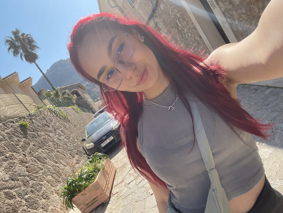 A selfie-style portrait captures a young female with long, dyed red hair and clear-frame eyeglasses, smiling subtly at the viewer. She wears a grey ribbed crop top and a silver-colored necklace with a heart pendant. A light-colored strap, consistent with a bag, crosses her chest. The background depicts a street scene in Valldemossa, Spain, under bright daylight. A textured stone wall runs along the left side, featuring a planter with green foliage and climbing vines. A dark-colored Ford vehicle, license plate '7557 HKT' partially visible, is parked on the paved street behind the subject. Traditional buildings with tiled roofs and a palm tree are visible against a clear blue sky. Mountains are discernible in the distant background. Bright sunlight illuminates the scene, creating a halo effect around the subject's hair and causing a lens flare in the upper right quadrant of the image.