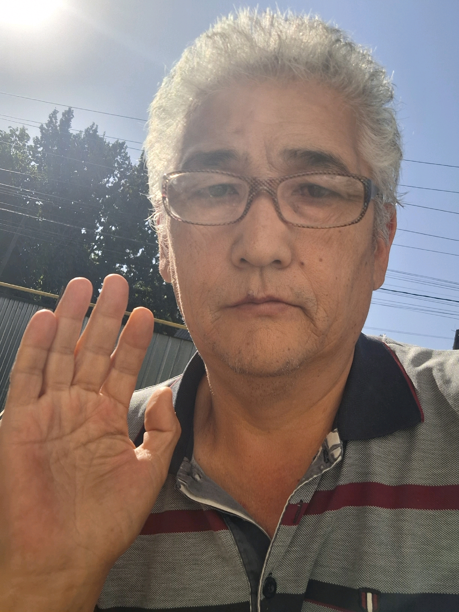A man with graying hair, wearing glasses and a collared shirt, holds his left hand up in front of his face. He is outdoors, with a bright sky and some trees visible in the background. The image is likely a selfie, as the camera appears to be held by the man. The scene suggests a casual, everyday moment. There is no specific information to determine the location within the city of Almaty, Kazakhstan.