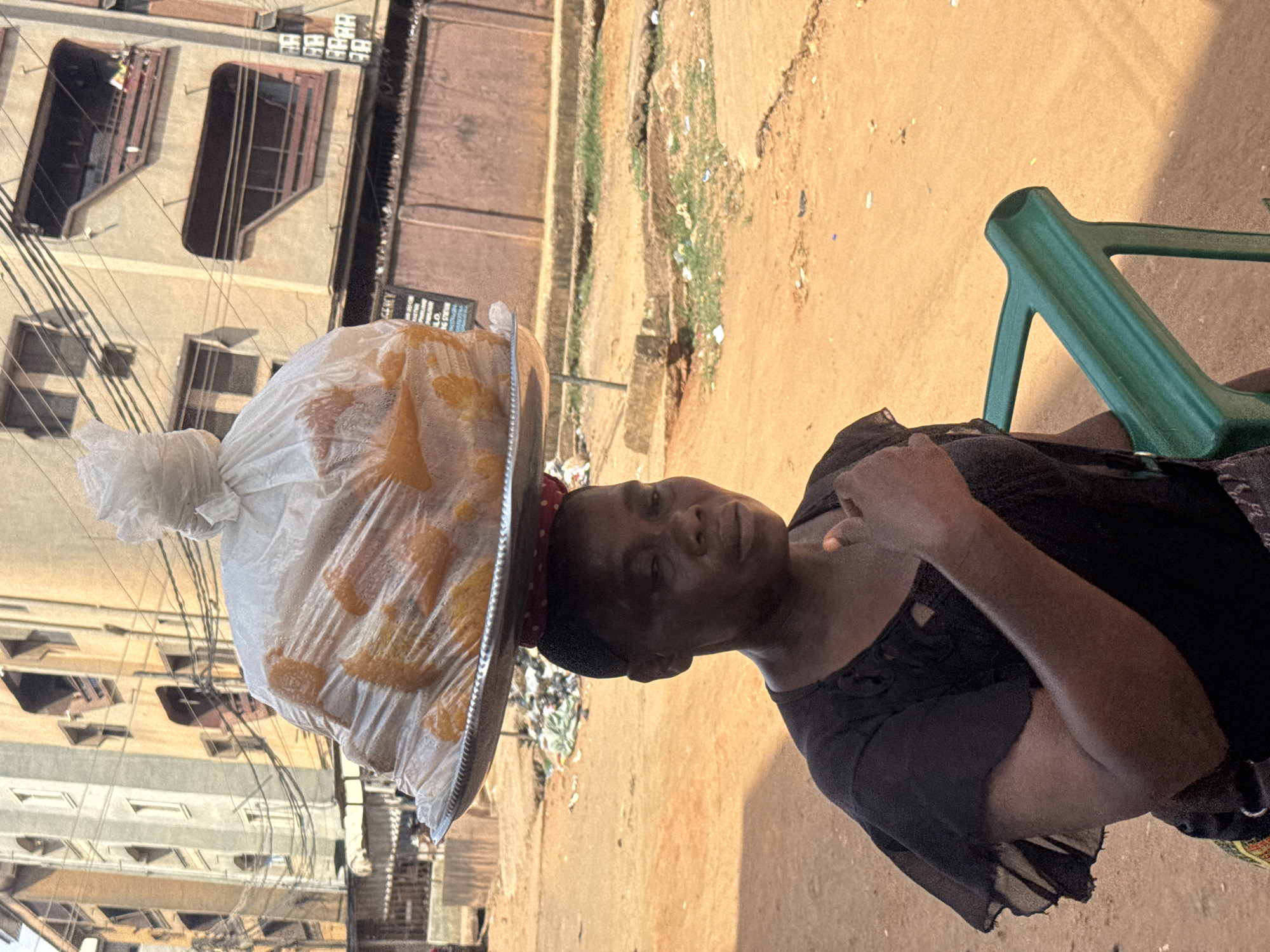 An outdoor, eye-level shot in Onitsha, Nigeria, shows a woman balancing a large tray of goods on her head while looking directly at the viewer. The woman, who has dark skin, is wearing a dark top or dress and a red and white polka-dotted head covering underneath a large, round, metallic tray. On the tray, a clear plastic bag is filled with numerous orange/yellow items, possibly fried snacks or baked goods, and tied securely at the top.
The setting appears to be an urban or semi-urban area. In the background, there are multi-story buildings with various windows and balconies, some with dark, recessed frames. Numerous electrical wires or clotheslines crisscross in front of these structures. The ground is unpaved, consisting of light brown earth or sand, with sparse patches of green vegetation and scattered debris. To the woman's right, a green plastic chair is partially visible, suggesting she might be resting or waiting.
The time of day is daylight, indicated by clear shadows and bright illumination, likely late morning or early afternoon. The weather appears sunny and clear. The woman's expression is neutral and calm. While some blurry text is present on a distant building in the background, possibly "BUSINESS LO" or similar, it is largely illegible. The image captures a common scene of street vending or hawking in the region.