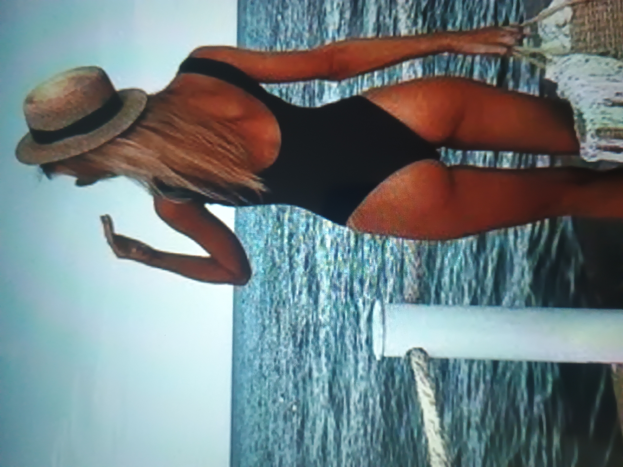 A person is captured from the back, standing on a pier or dock, facing a large body of water under a clear sky in the city of Unknown, Unknown. The individual has long, light-colored hair and is wearing a dark one-piece swimsuit and a light-colored straw hat with a dark band. Their right arm is raised, with the hand positioned near their head. In their left hand, they hold a light-colored woven bag with a white cloth draped over it. To the left of the person, a white cylindrical post is visible, with a thick, light-colored rope secured to it. The background consists of a vast expanse of blue-green sea with ripples, meeting a bright, clear blue sky at the horizon.