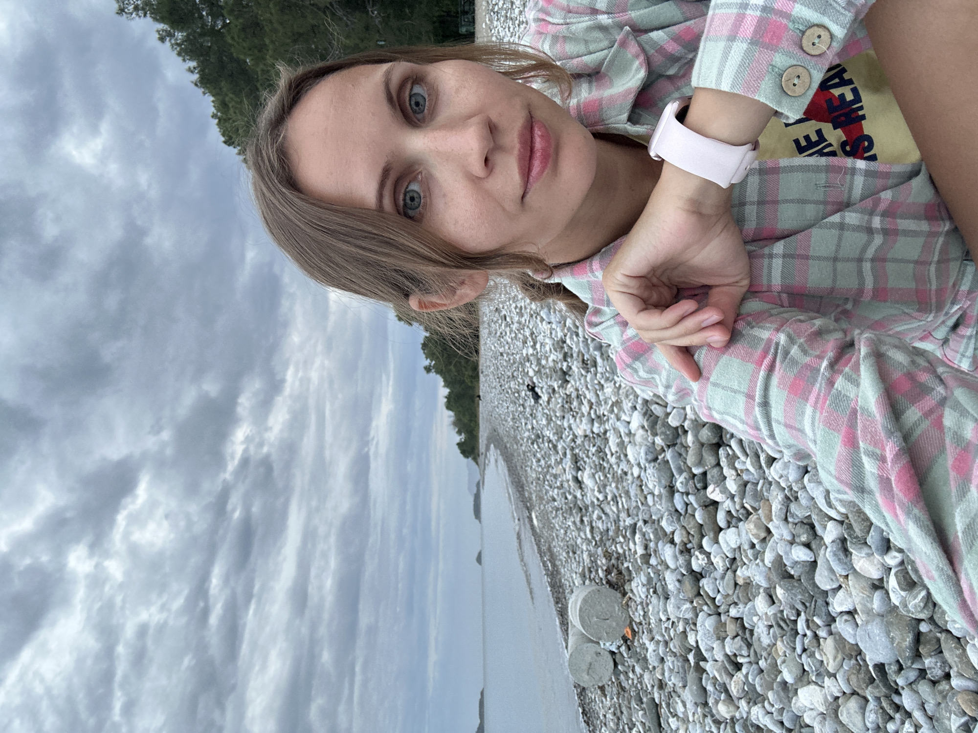 A young woman with light complexion, blue eyes, and light brown hair is positioned in a selfie on a rocky beach. She wears a pink and green plaid shirt and a light pink smartwatch. The background reveals a pebble beach, water, and cloudy sky, suggesting an overcast day. Two cylindrical concrete structures are visible on the beach near the water. The location is likely in Fethiye, Turkey, evidenced by the beach landscape.