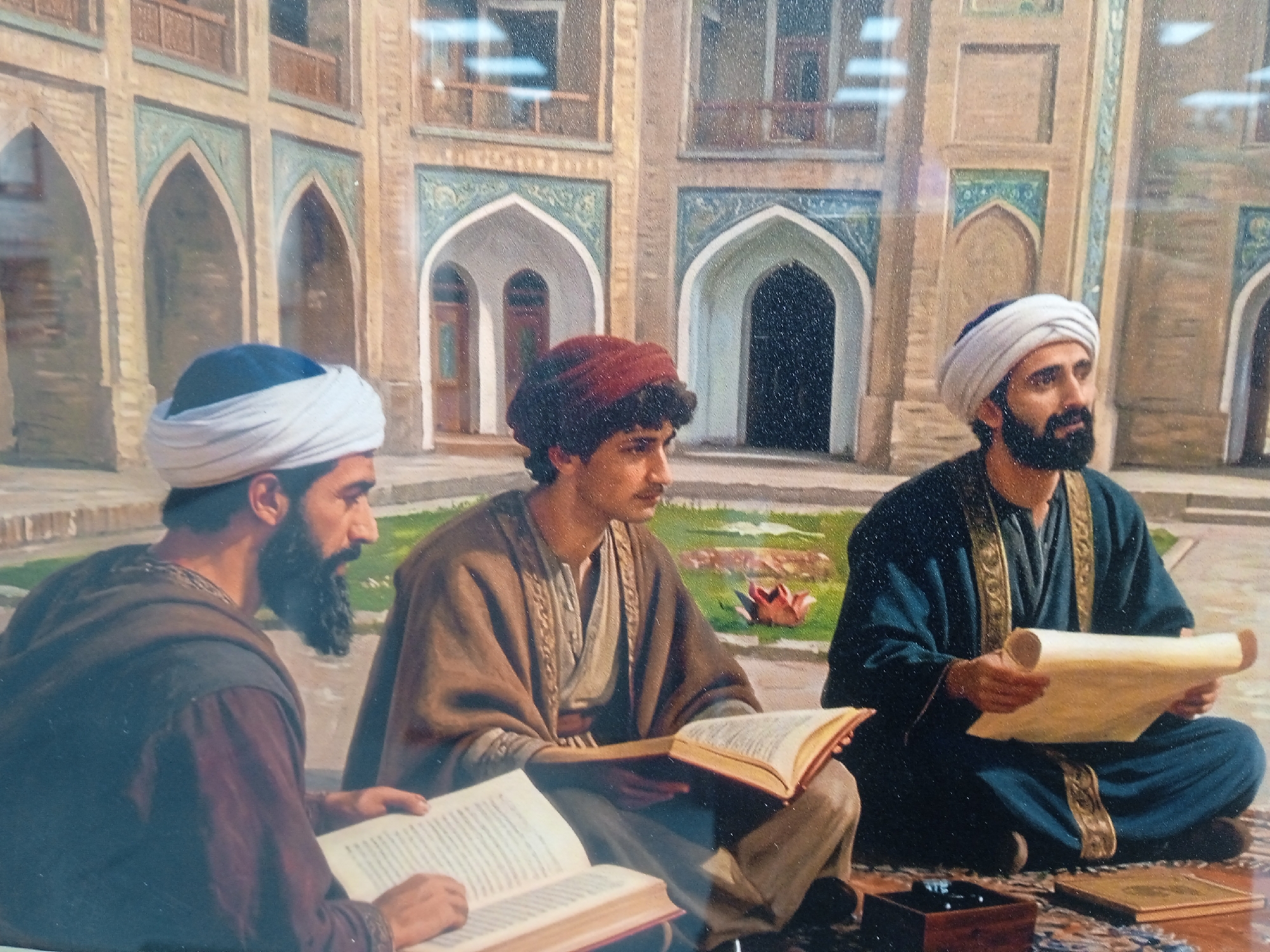 The image depicts a detailed painting of three men in traditional attire sitting in an open courtyard. The architecture suggests a historical Islamic setting with arched doorways and intricate tilework. The men are engaged in scholarly activities.
The man on the left, partially visible, wears a white turban and a dark robe. He is holding a large open book. The central figure, also in a white turban and a light brown cloak over a patterned garment, is reading an open book. The man on the right, sporting a white turban and a dark blue robe with golden trim, holds a rolled-up scroll. The ground is covered with a patterned rug, and a wooden box and another book are visible nearby. The background shows a building with multiple levels and ornate windows, characteristic of historical Middle Eastern or Central Asian architecture. The lighting suggests an outdoor, daytime setting.