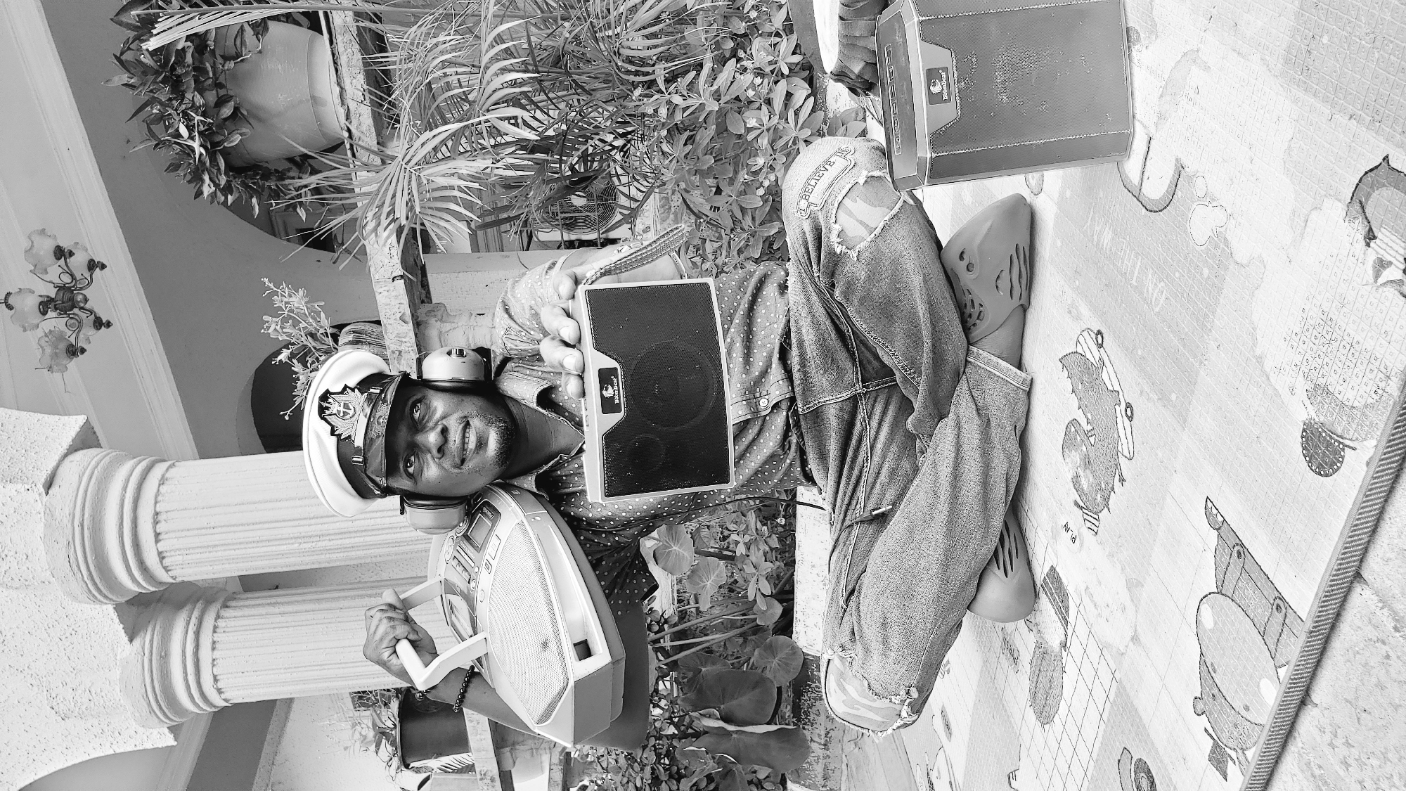 In Sabon Tasha, Nigeria, a Black male wearing a captain's hat and headphones is seated cross-legged on a patterned mat. He is holding a portable boombox in his left hand, resting it on his shoulder, and extends his right hand forward, holding a smaller speaker. He is dressed in a patterned shirt and ripped jeans. To his right, another speaker sits on the mat. The background features architectural elements, including pillars and a railing, with lush green plants and a chandelier visible. The scene is captured in black and white.