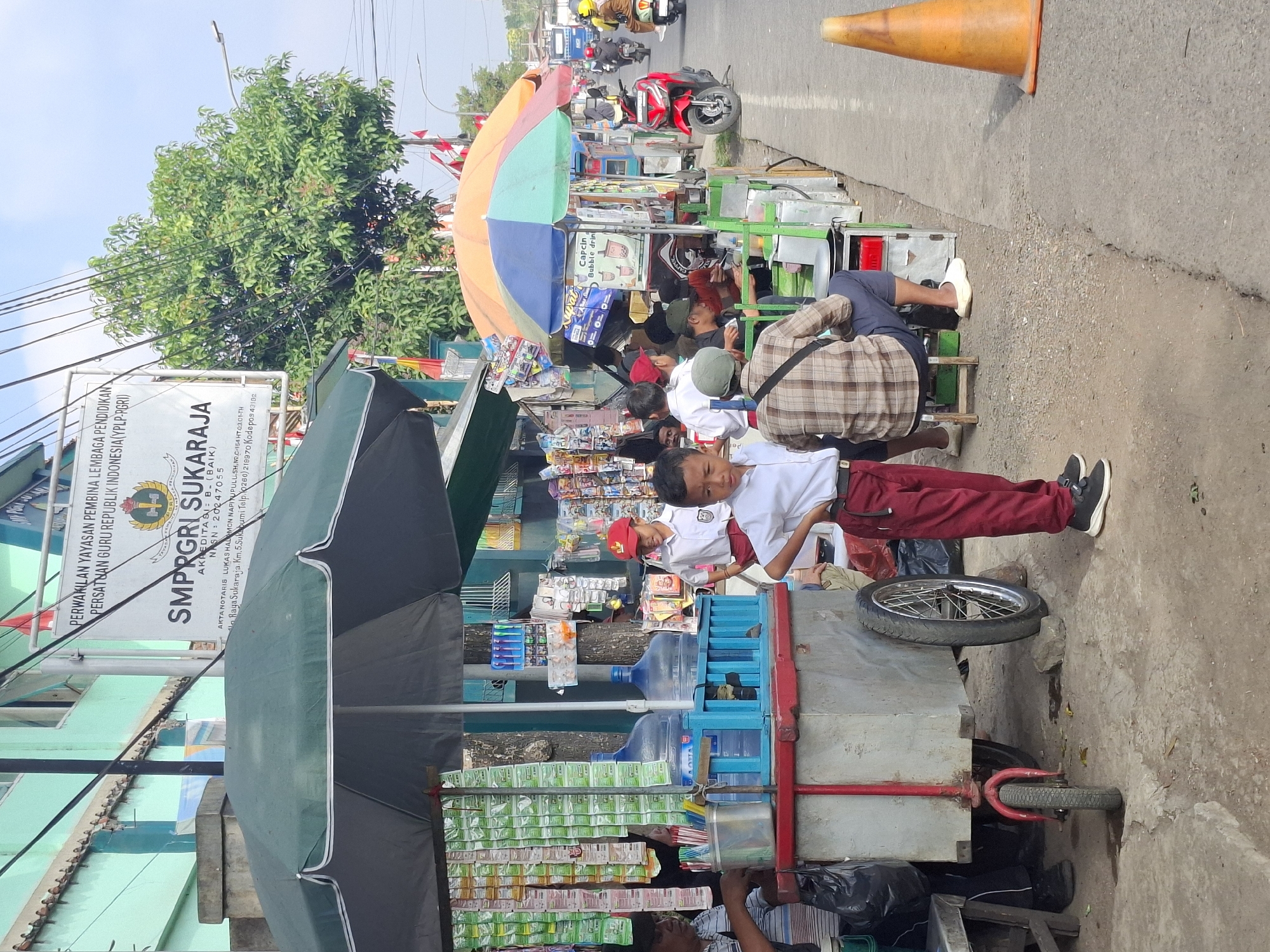 In Sukaraja, Indonesia, a street scene unfolds with a focus on a roadside vendor and pedestrians, including schoolchildren. The environment is bustling, with a paved road running parallel to a sidewalk or dirt path where various stalls are set up. A prominent green umbrella shades a cart displaying rows of packaged snacks and drinks. Two large blue water jugs are visible behind the cart.
Several individuals are present. A young boy in a white shirt and red pants stands with his hands on his hips, facing the camera. Another boy, also in a school uniform, stands nearby, looking towards the right. An older man in a striped shirt is seated behind the cart, seemingly attending to the stall. Further down the street, several people are gathered around other vendors, one of whom is preparing food. A man in a plaid shirt and blue shorts is seated on a stool, facing away from the camera.
The background reveals more of the urban setting, including a building with turquoise walls and windows. A sign above the vendor area indicates "SMPRGR SUKARAJA," suggesting the presence of a junior high school. Traffic, including motorcycles, is visible on the road, with one red motorcycle parked prominently. An orange traffic cone stands on the side of the road. The overall atmosphere is that of a typical Indonesian street market or roadside commercial area.